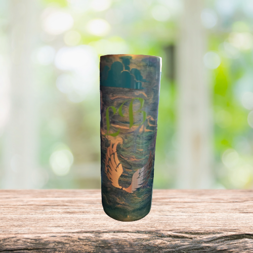 A ceramic mug with a landscape painting, featuring mountains, sky, and a hand reaching out, placed on a wooden surface with a blurred outdoor background.