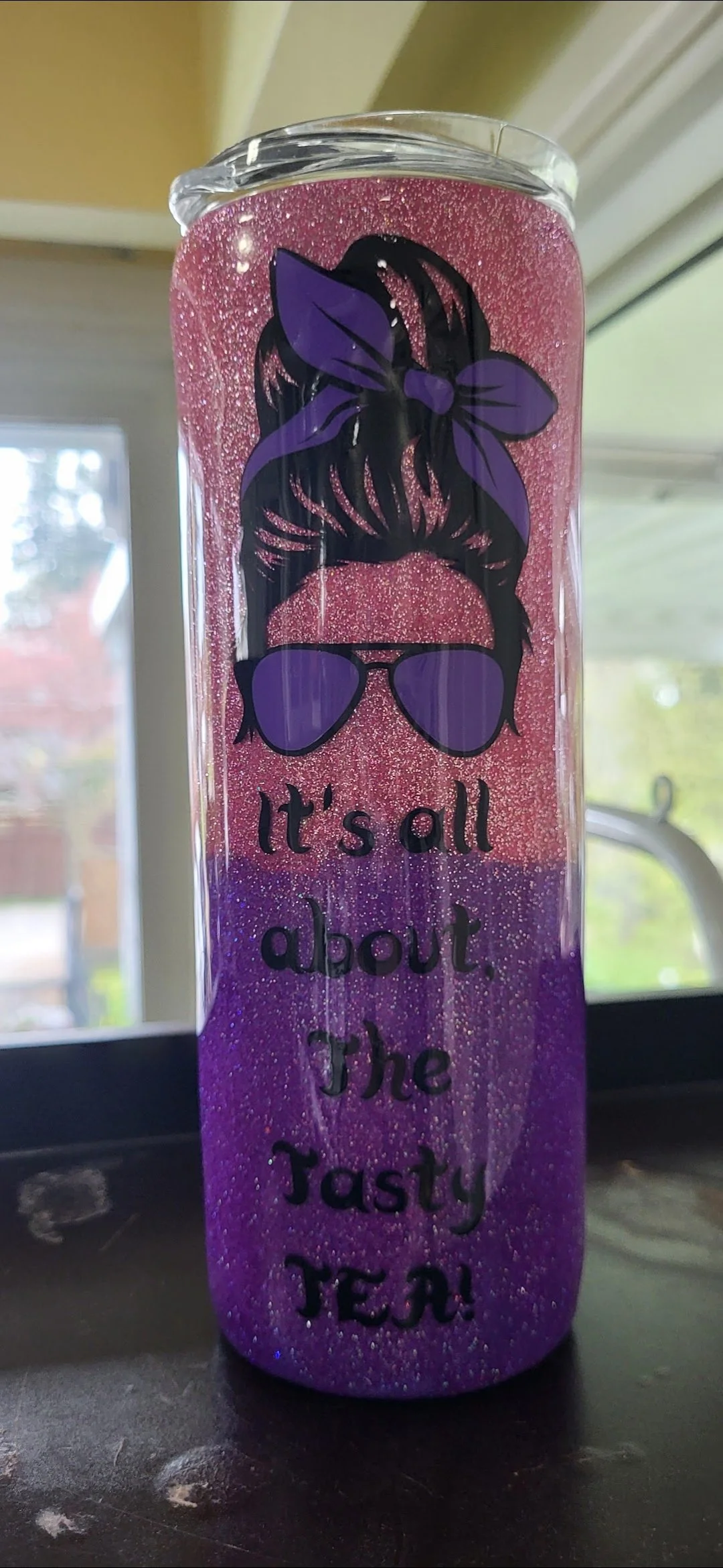 A glittery tumbler cup with a graphic of a woman's hair in a bun, wearing purple sunglasses and a purple headband, and the text 'It's all about the Tasty TEA!' written in black.