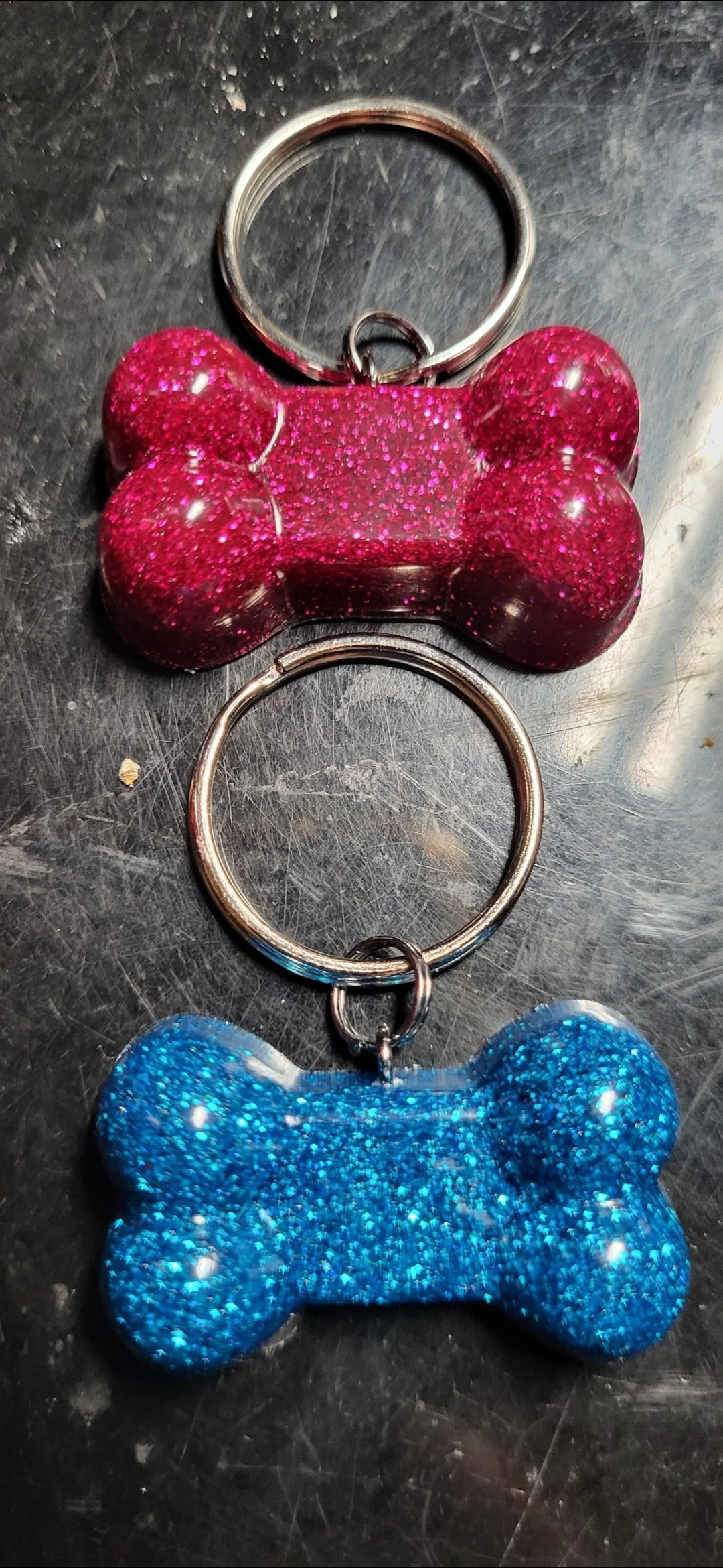 Two glittery dog bone-shaped keychains, one pink and one blue, on a metallic surface.