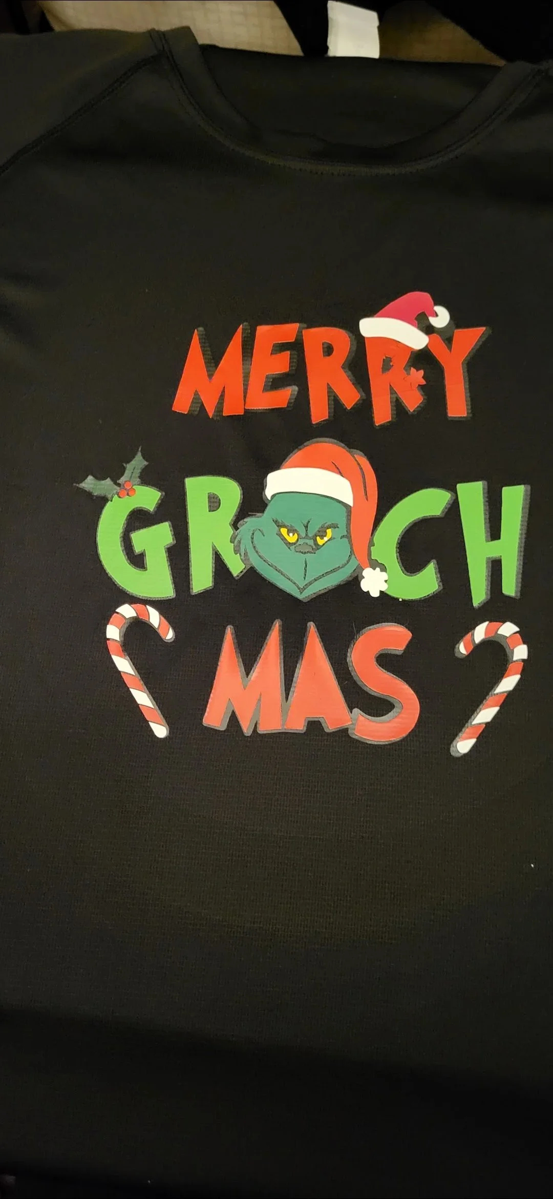A black shirt with a festive, Grinch-themed holiday design that reads "Merry Grinchmas" with a Grinch face wearing a Santa hat, candy canes, and holly decorations.