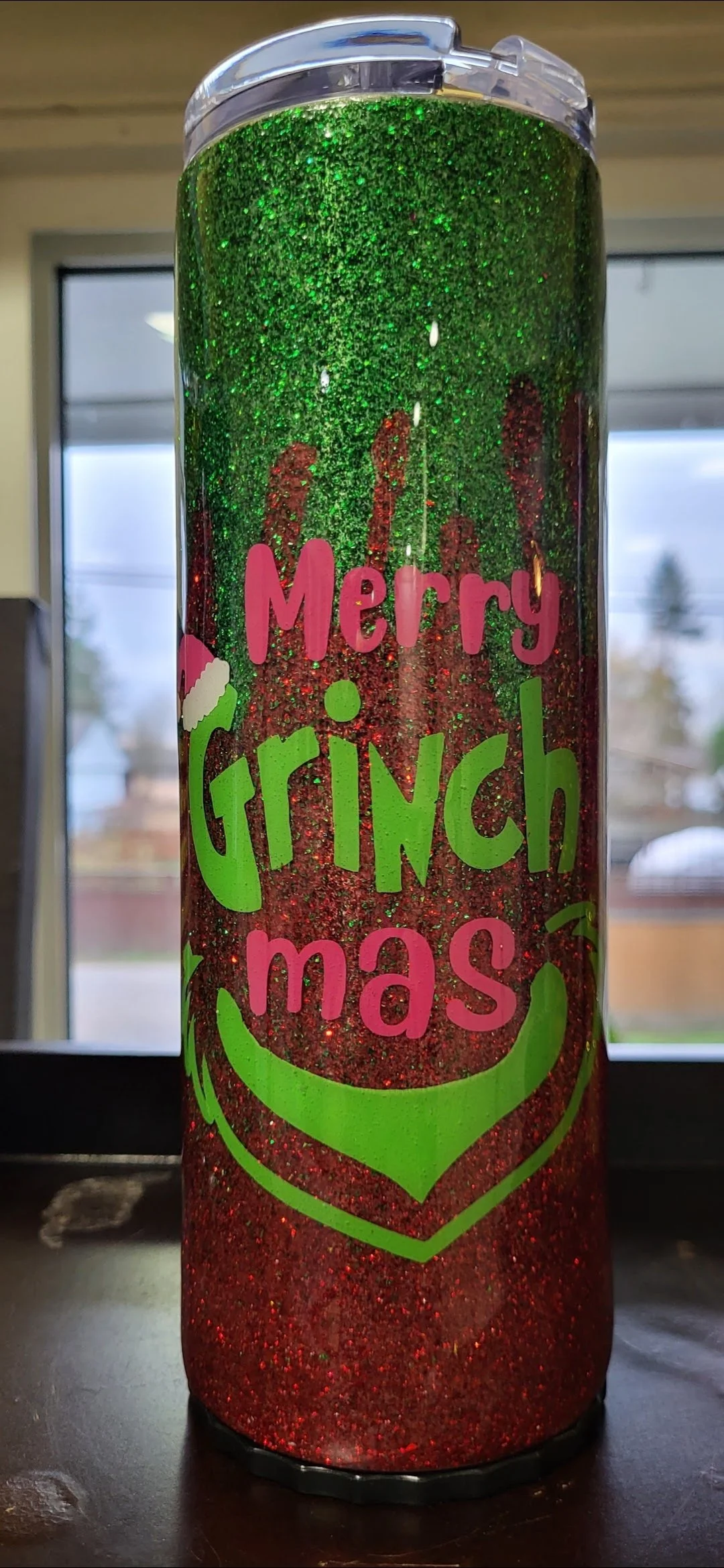 A large holiday-themed tumbler decorated with red and green glitter, displaying the message "Merry Grinch-mas" in bright pink and green lettering, with a smiley face graphic.