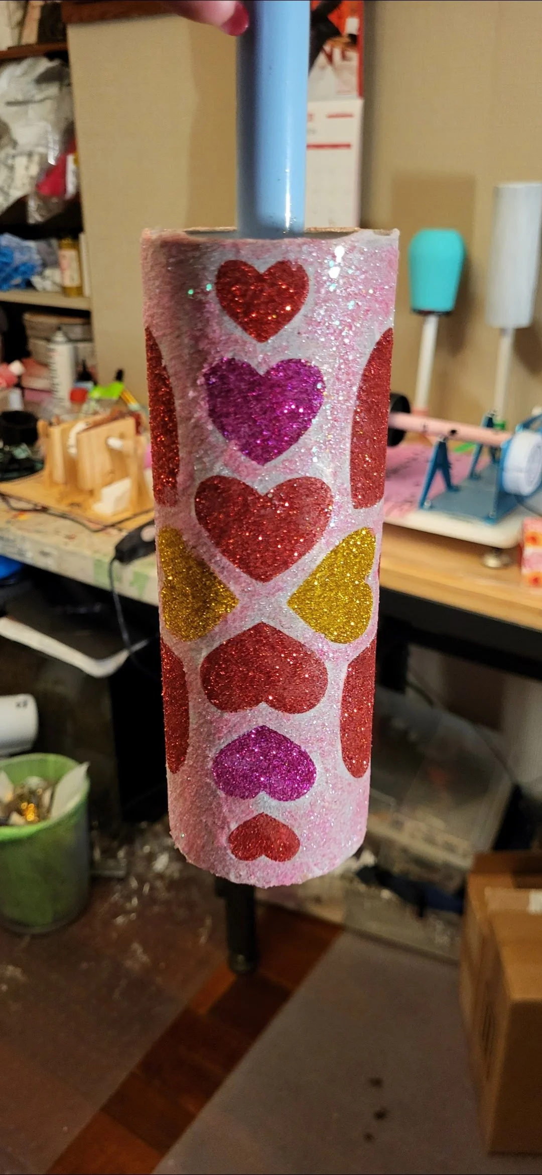Decorated cardboard tube with pink glitter and heart designs in red, purple, and yellow, hanging from a blue pipe, with a cluttered workshop background.