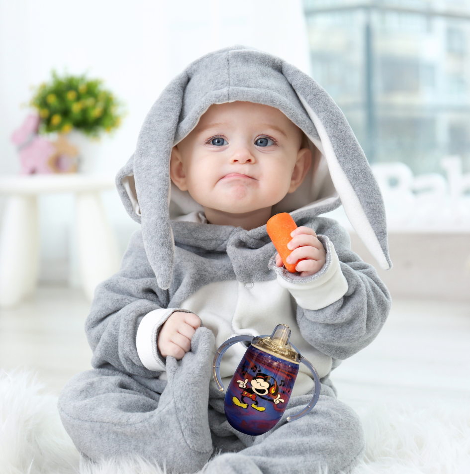 Baby dressed in a grey bunny costume holding a carrot and a sippy cup, sitting indoors near a window with a blurred background of a plant and a pink decorative item.