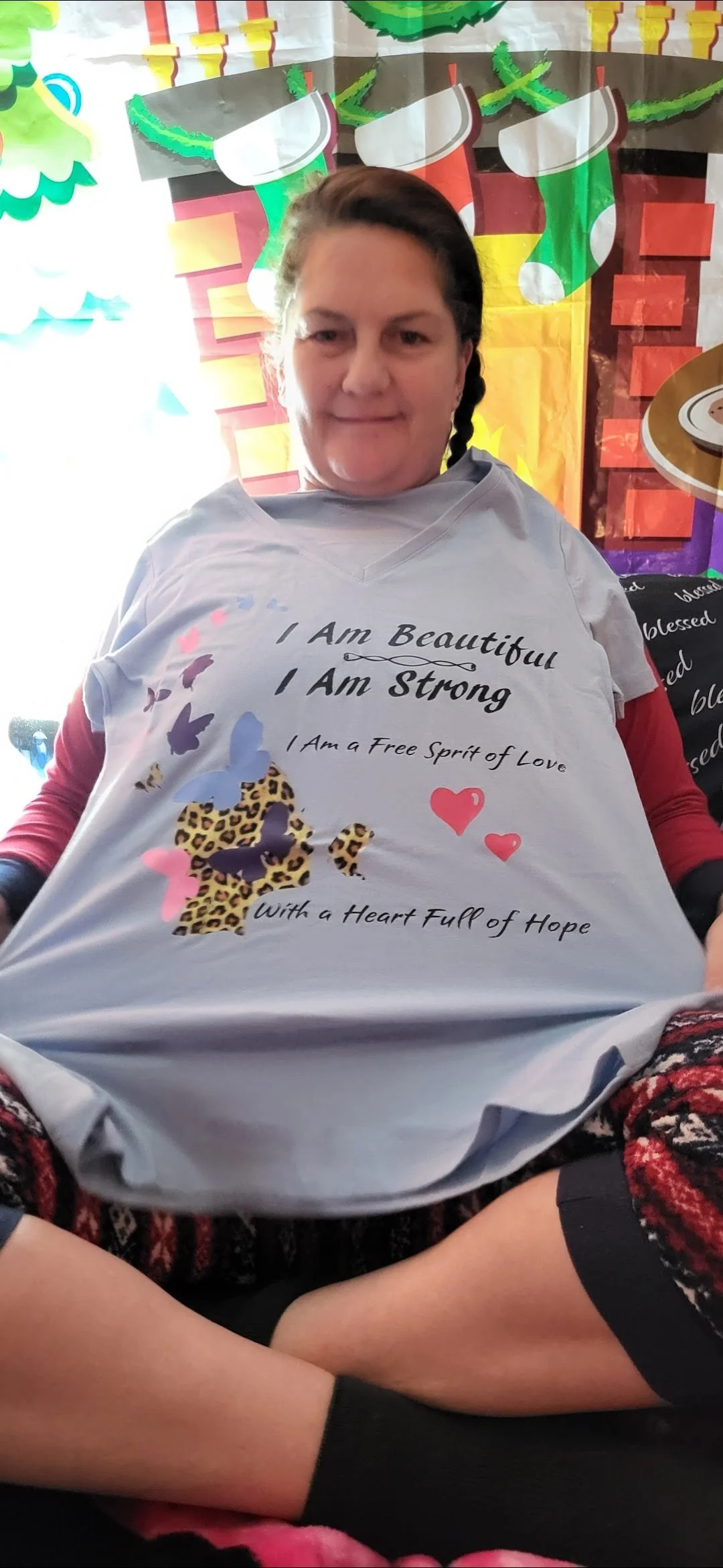 A woman sitting in front of a colorful holiday-themed backdrop wearing a light-colored shirt with positive affirmations and butterfly and animal print designs.