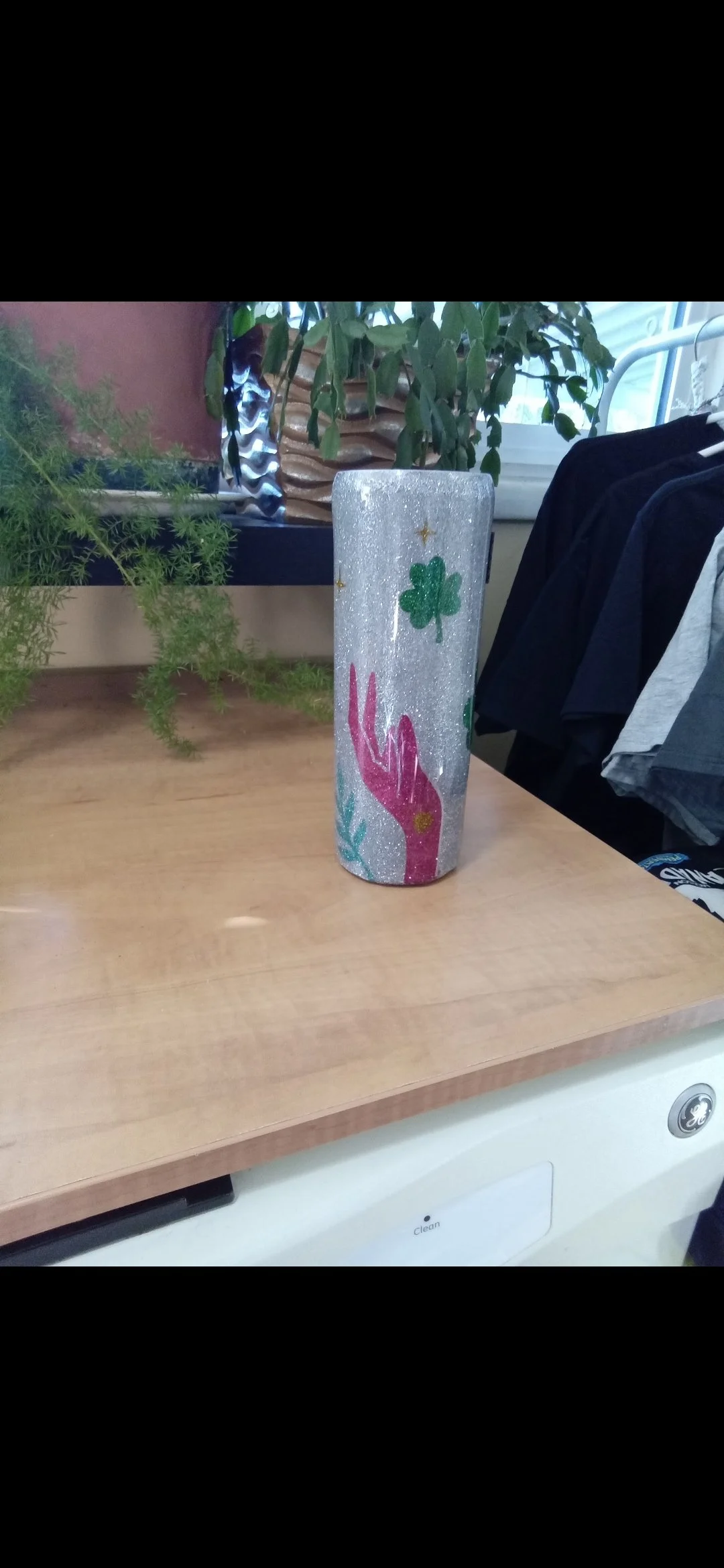 Decorative cylindrical vase with glitter and hand-painted green shamrocks, pink hand, and other colorful designs, placed on a wooden surface with potted plants and clothing nearby.