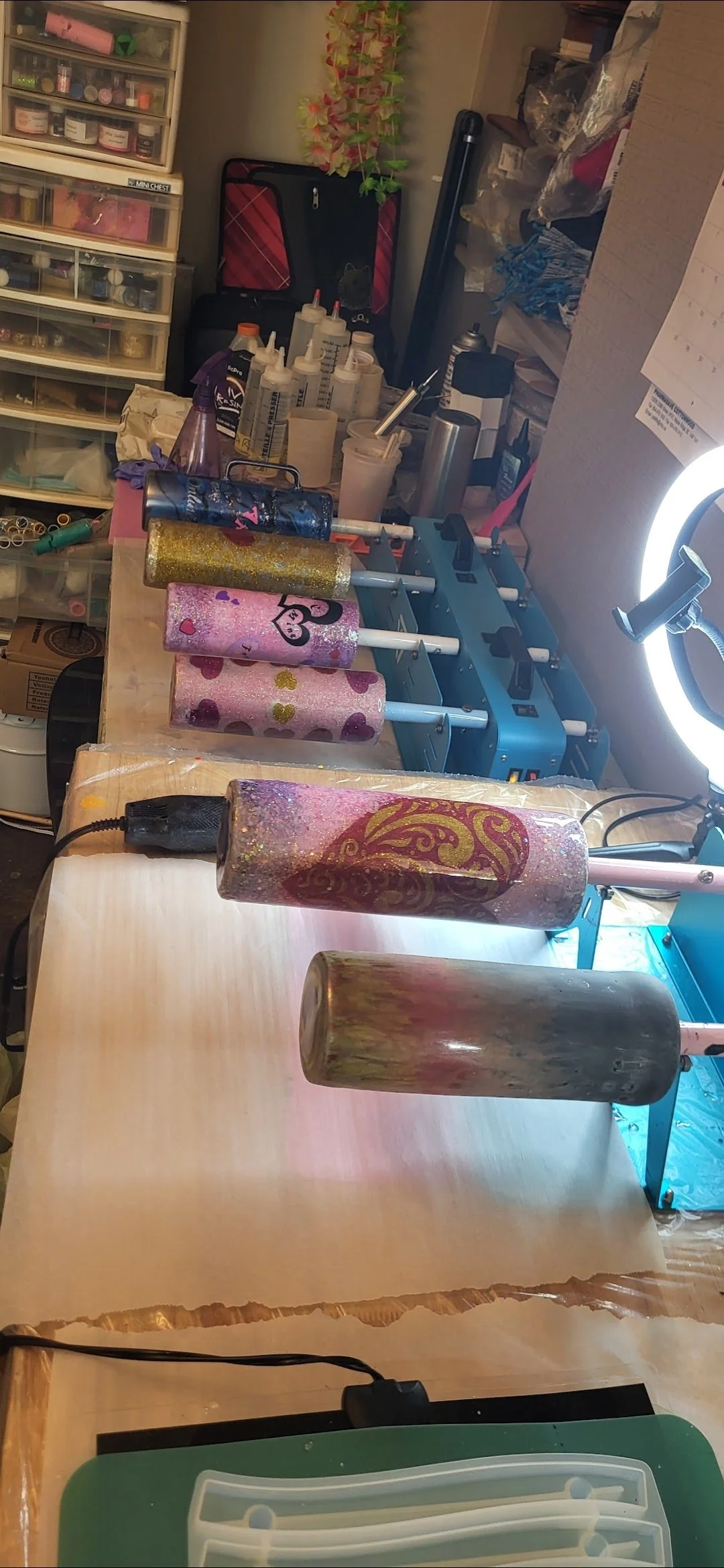 A workshop with glittered tumblers in various stages of decoration on a wooden surface, surrounded by craft supplies, tools, and storage drawers.