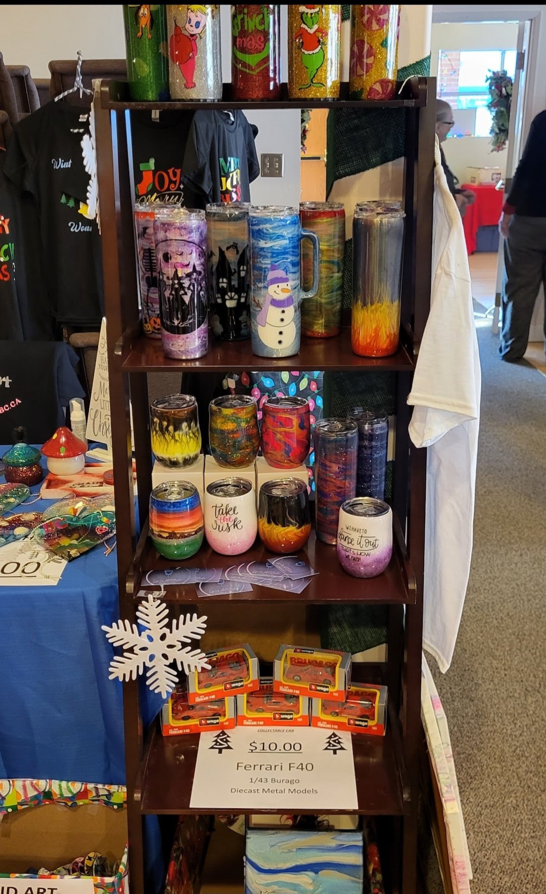 Display shelf with colorful Christmas-themed tumblers, mugs, and decorative items, including toy cars and handmade art pieces.
