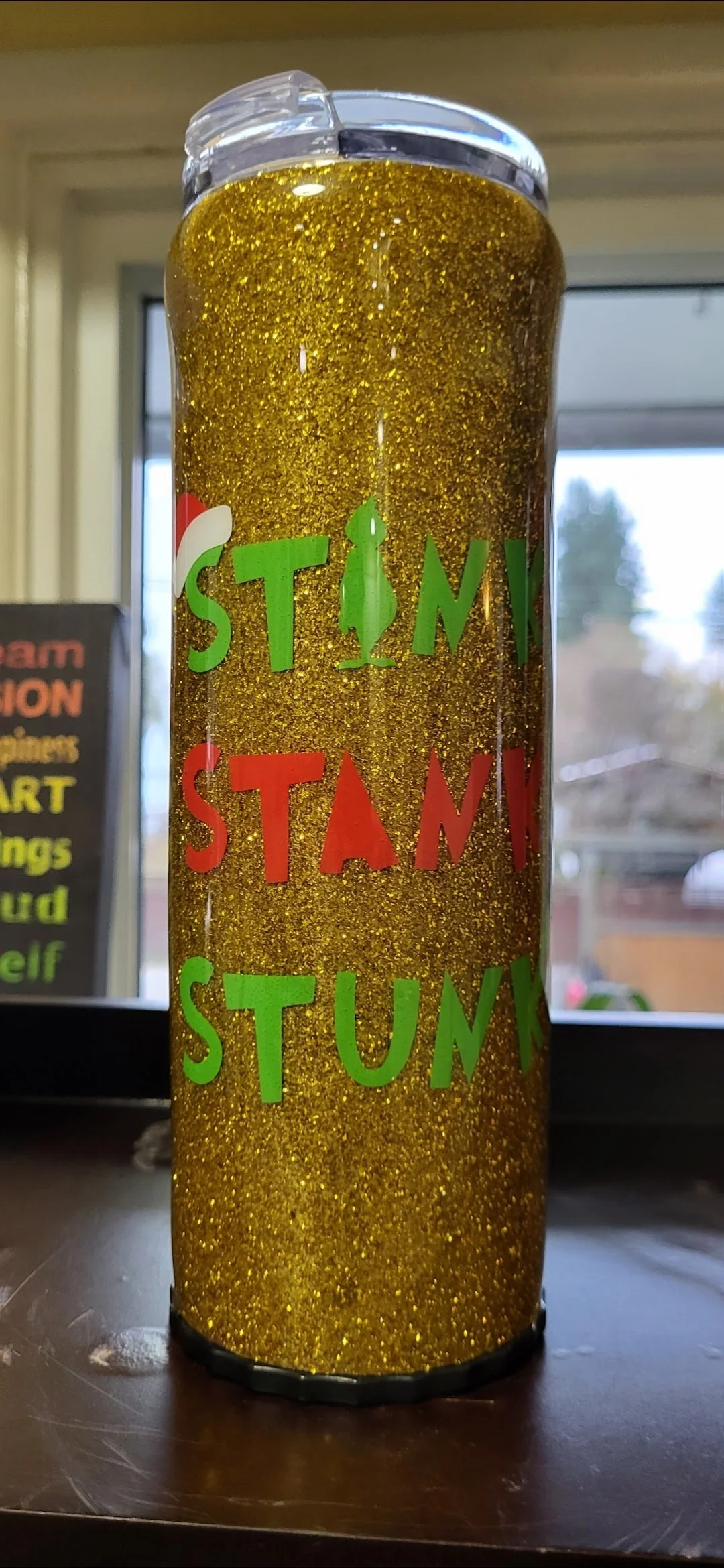 A tall cylindrical tumbler decorated with gold glitter and colorful stickers spelling out 'STAM' in green and red letters, with additional small decorative elements. It is placed on a dark surface with a window in the background.