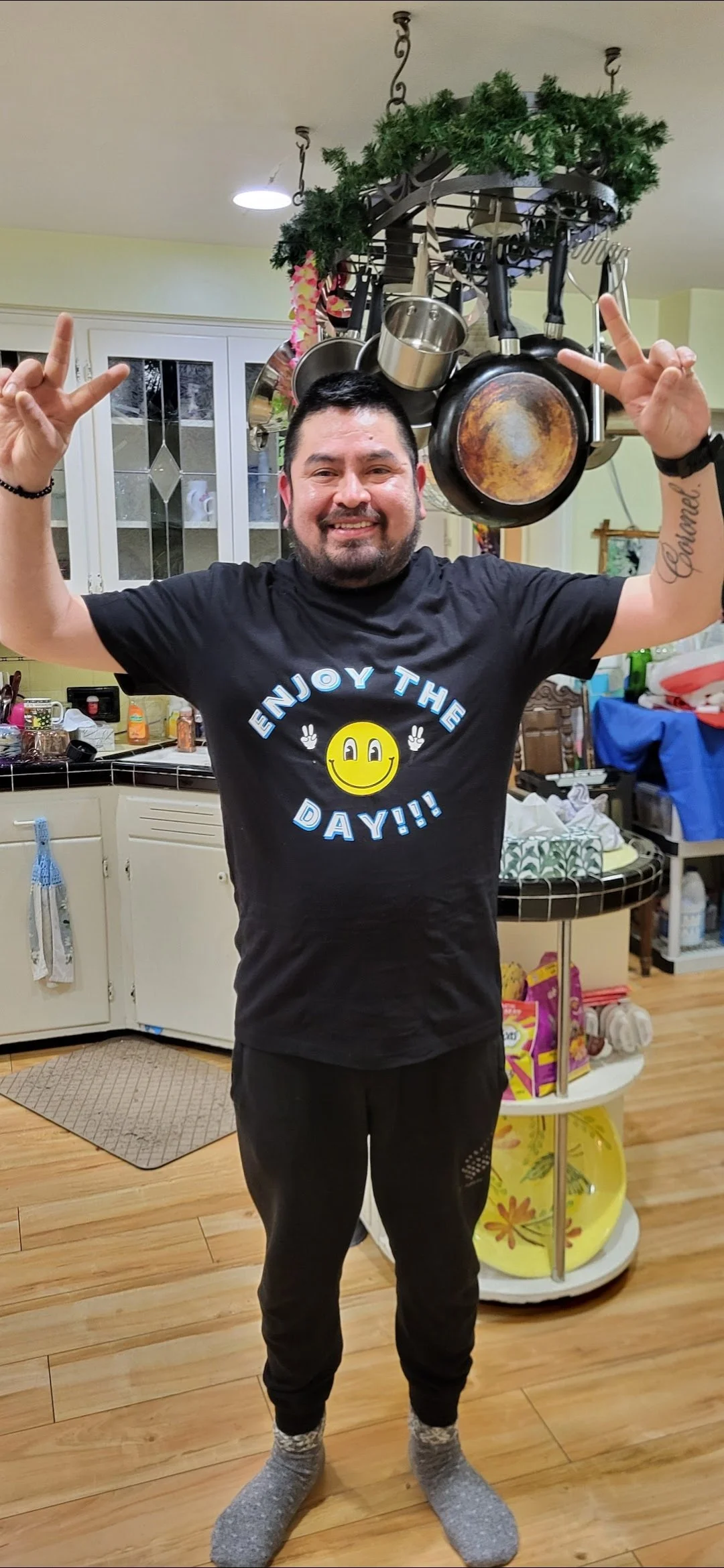 Smiling man standing in a kitchen, raising both hands with peace signs, wearing a black shirt with 'Enjoy the Day!!' and a yellow smiley face, with pots hanging from a ceiling rack behind him.