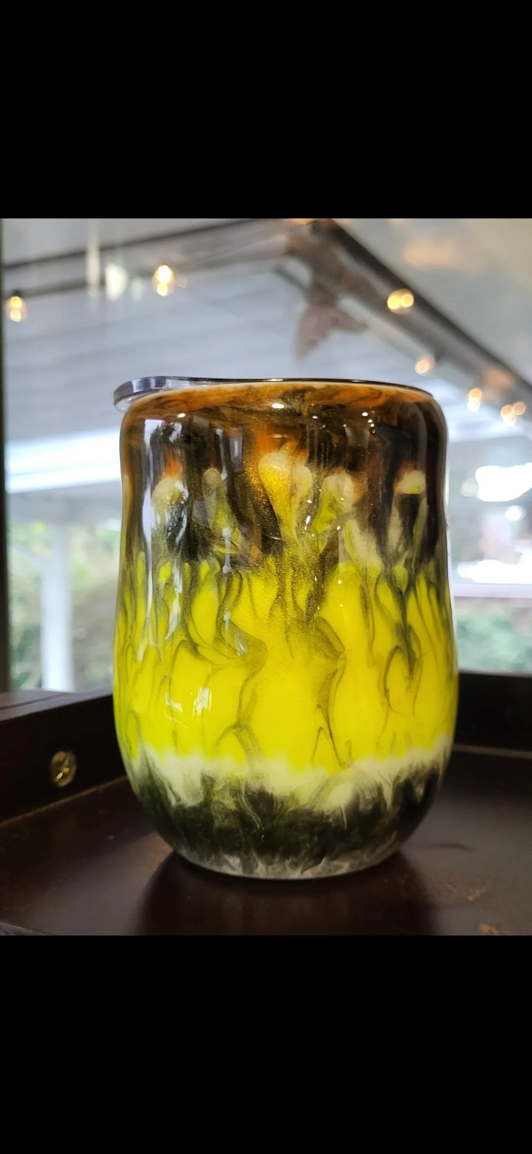 Colorful glass vase with yellow, black, and white swirling patterns, sitting on a dark surface in a room with string lights.