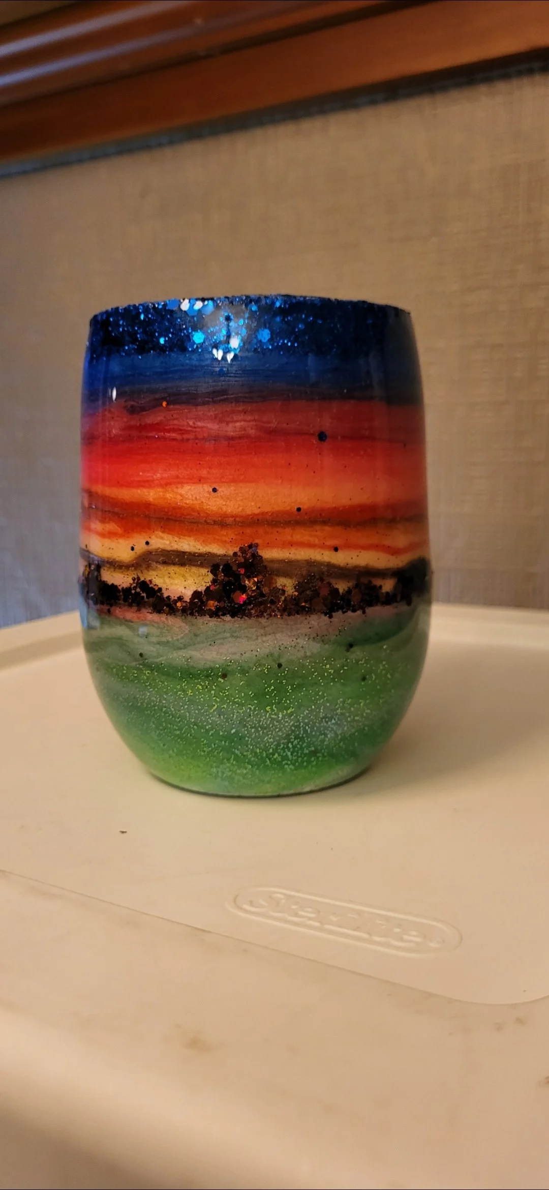 A colorful, hand-painted glass with a sunset landscape scene, featuring a dark blue sky with glitter, orange and red hues for sunset, black details for trees or ground, and green grass at the bottom, placed on a beige surface.