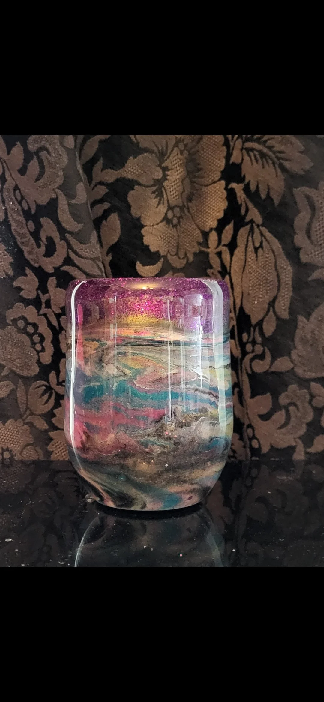 A colorful, marbled glass candle holder with a lit candle inside, resting on a reflective black surface with a floral patterned fabric background.