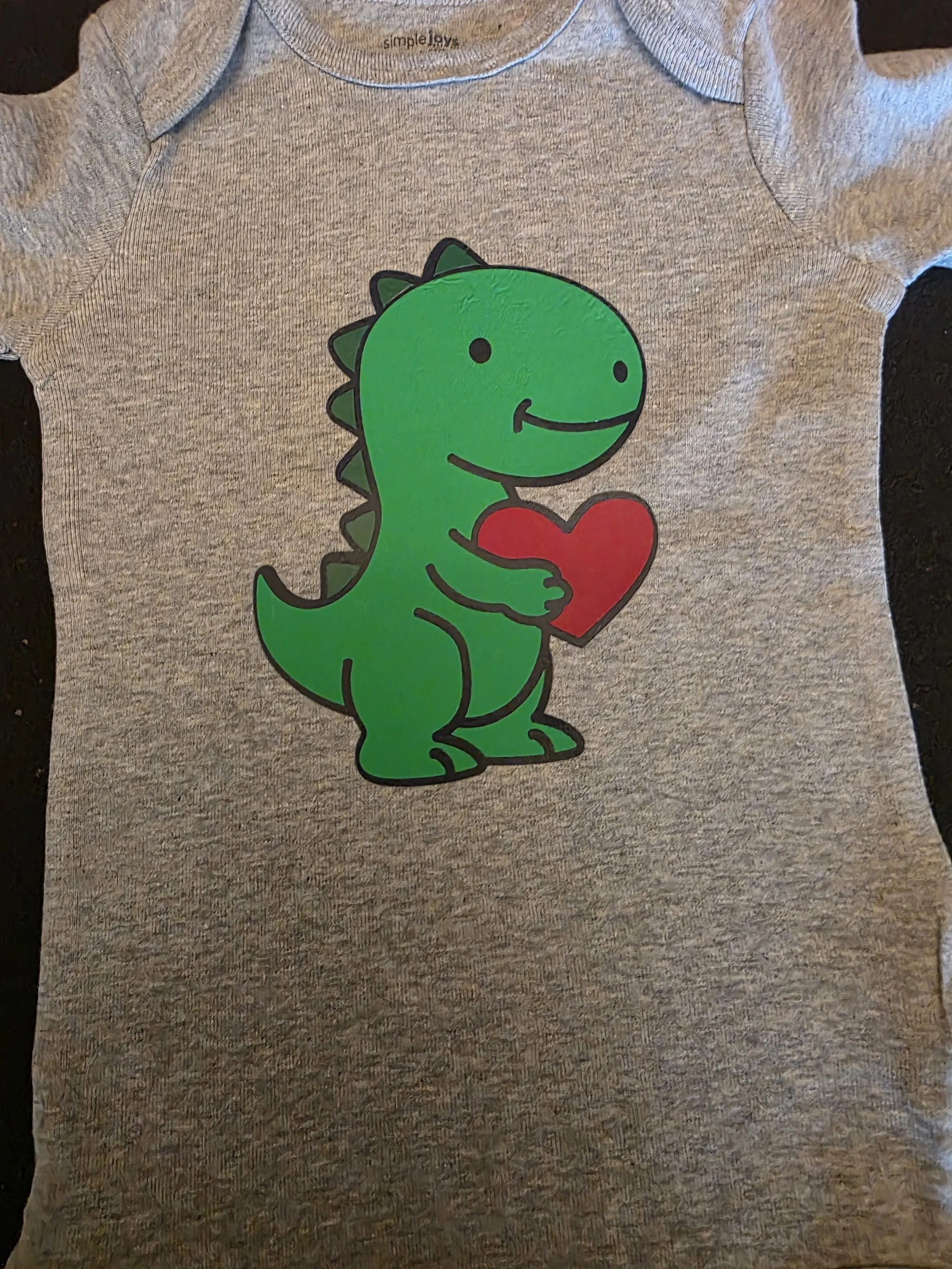 Gray T-shirt with a cartoon green dinosaur holding a red heart, smiling.