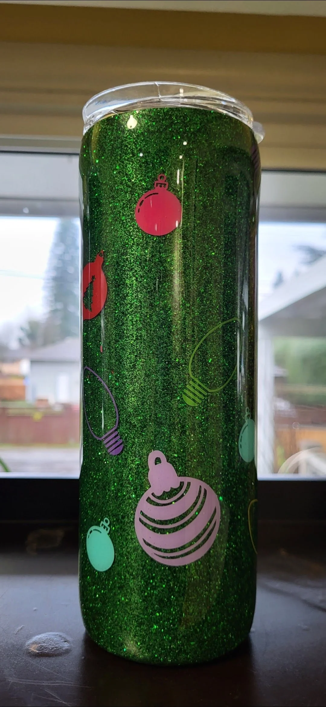 A clear plastic tumbler with green glitter and holiday-themed ornaments and lights printed on it, sitting on a dark surface with a window and houses in the background.