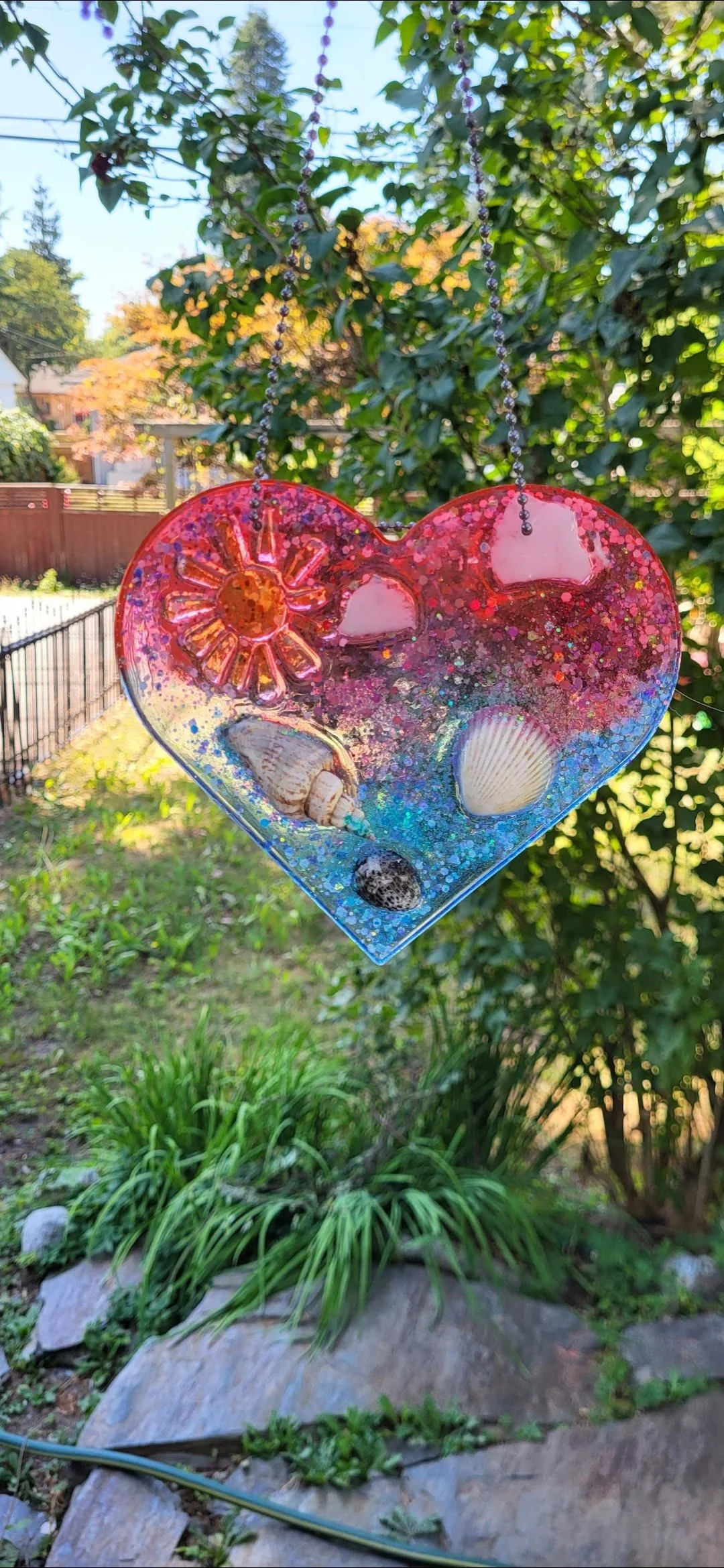 Colorful resin heart-shaped ornament with seashells and glitter, hanging outdoors in a garden.
