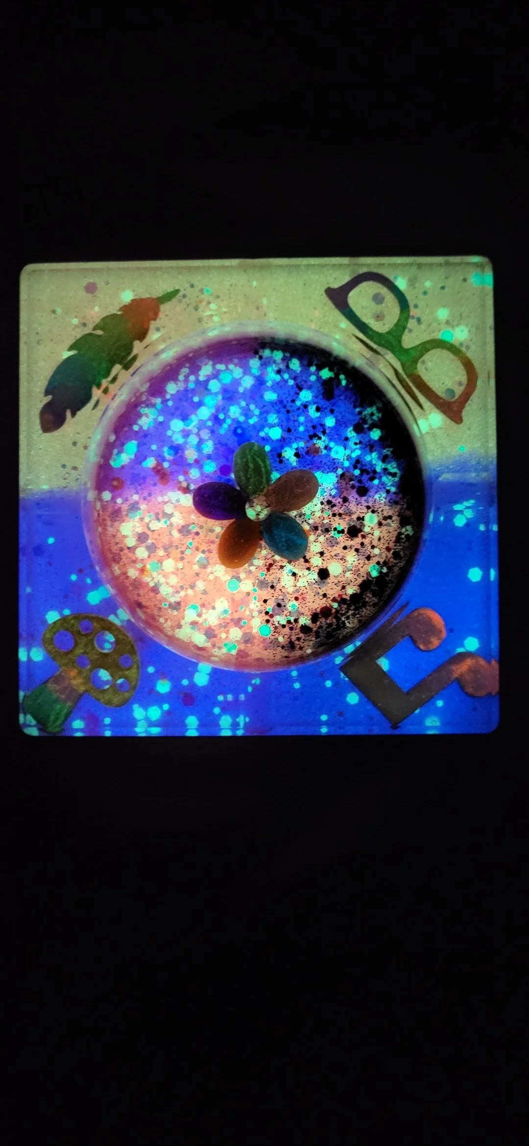 A small square, glowing, rainbow-hued slime container with a flower-shaped decoration inside. The container has colorful, cartoon-like illustrations of a beetle, a mushroom, and glasses around its edges.