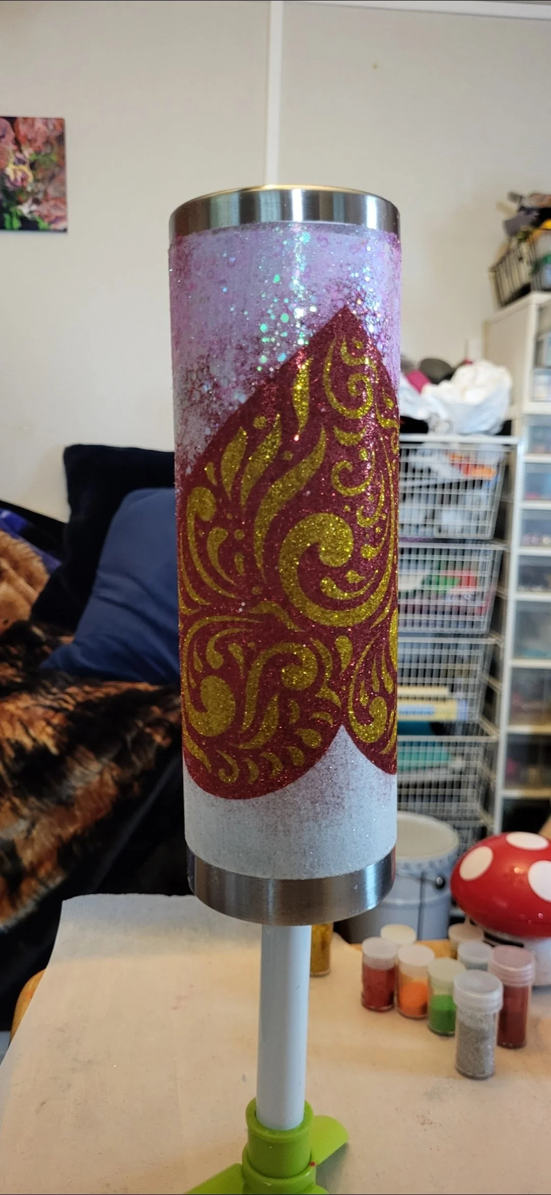 Decorative cylindrical object with a purple, white, and gold ornate design, mounted on a white stand with a green base. The background shows a cluttered room with storage drawers, clothes, and small containers of craft supplies.