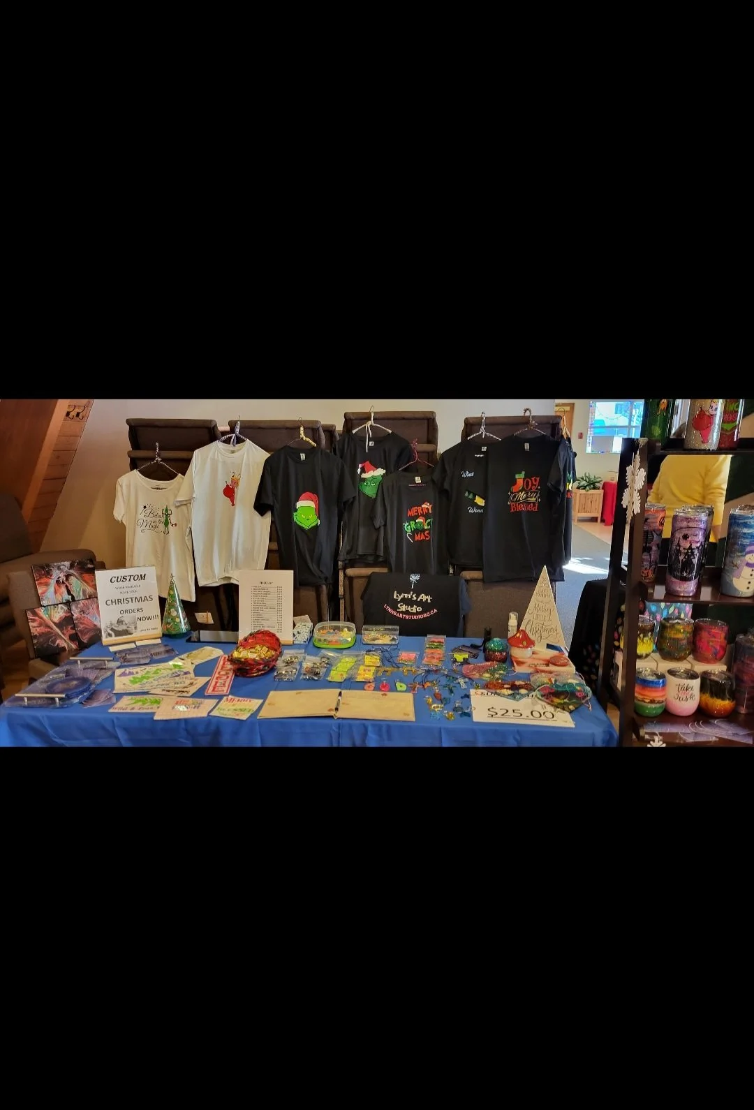 Table setup at a Christmas craft fair with custom Christmas items, including T-shirts with holiday designs, jewelry, and decorated candles, with signs indicating prices and ordering information.