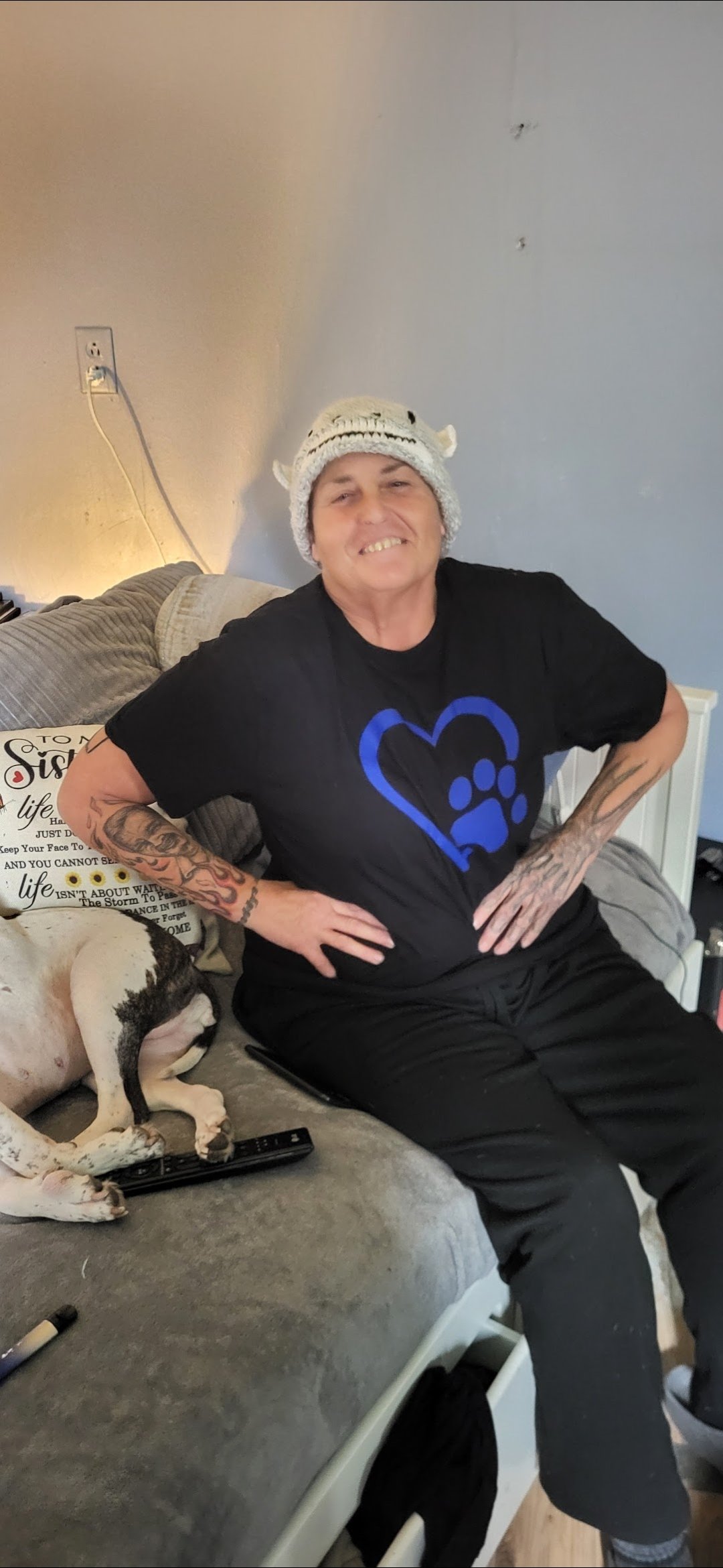 A person with tattoos on arms, wearing a black shirt with a blue heart and paw print, sitting on a couch with a dog lying beside them. The person is smiling and wearing a white knit hat with small ears.