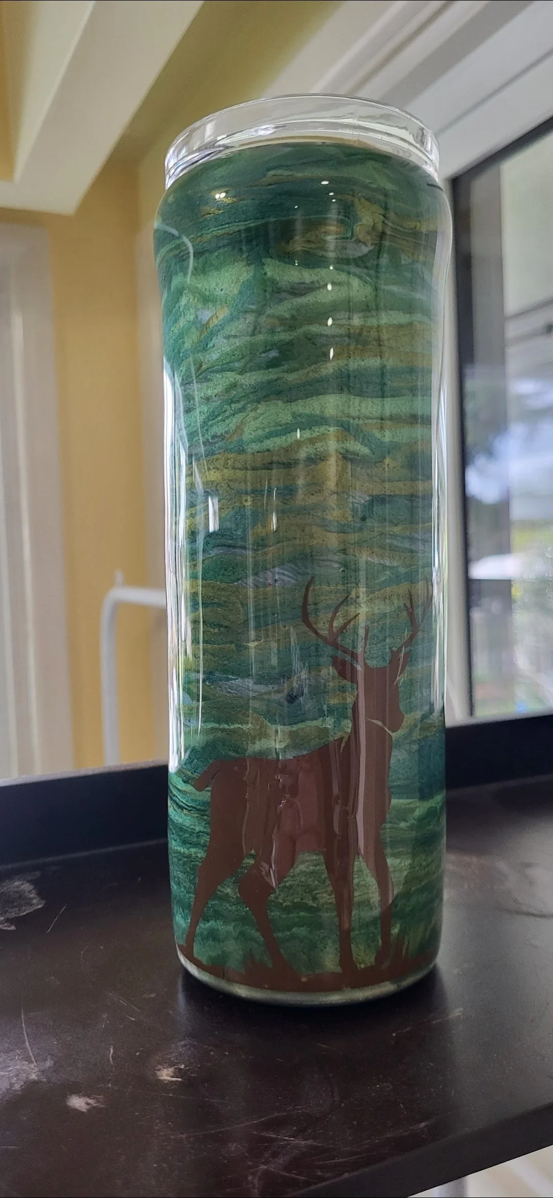 A cylindrical glass with a nature-themed design featuring a deer standing under a tree, with a background of green hills and sky.