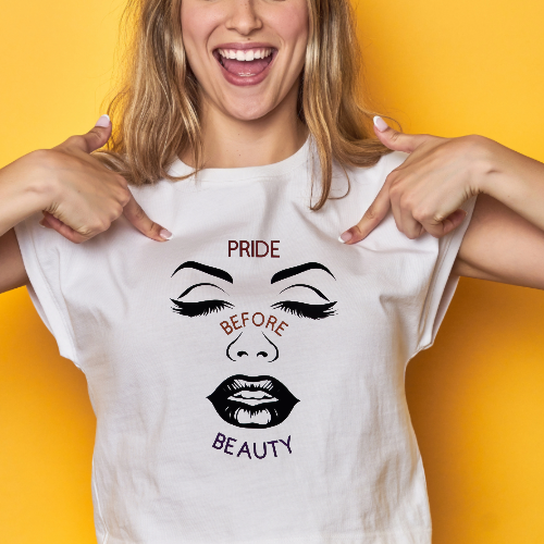 A smiling woman pointing at her white T-shirt with a graphic face design and the words 'PRIDE BEFORE BEAUTY' against a yellow background.