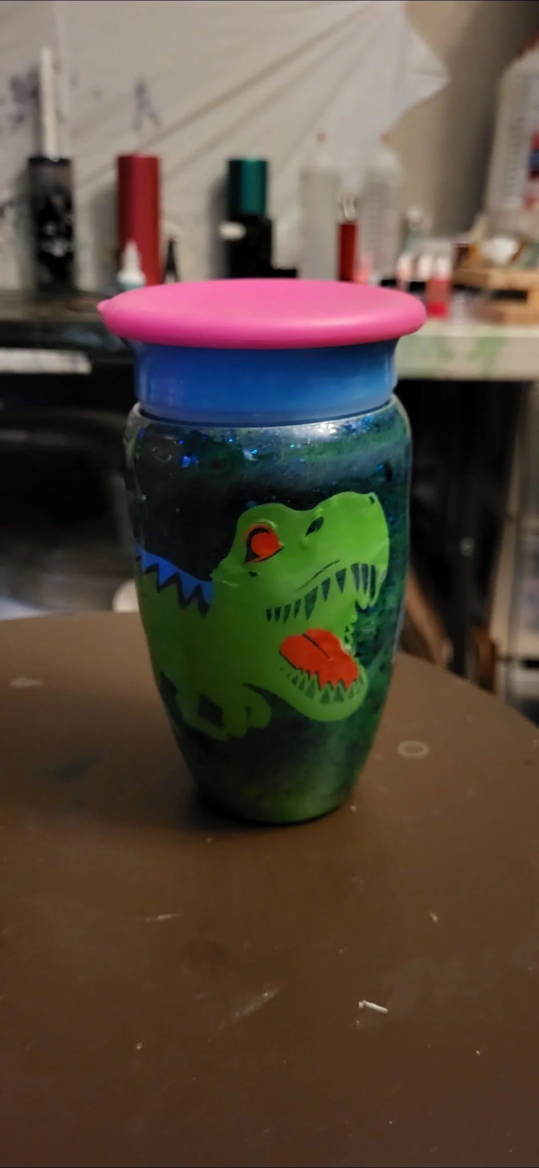 A decorated glass jar with a dinosaur illustration, topped with a pink lid, placed on a brown surface in a room with various items in the background.