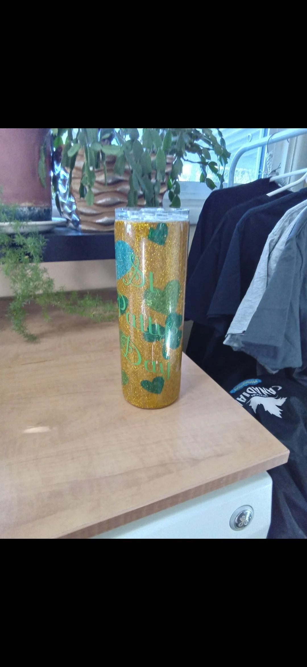 Gold glitter tumbler with green and blue heart designs and the words 'I Love Dad' in green cursive lettering, placed on a wooden surface with plants and clothes in the background.