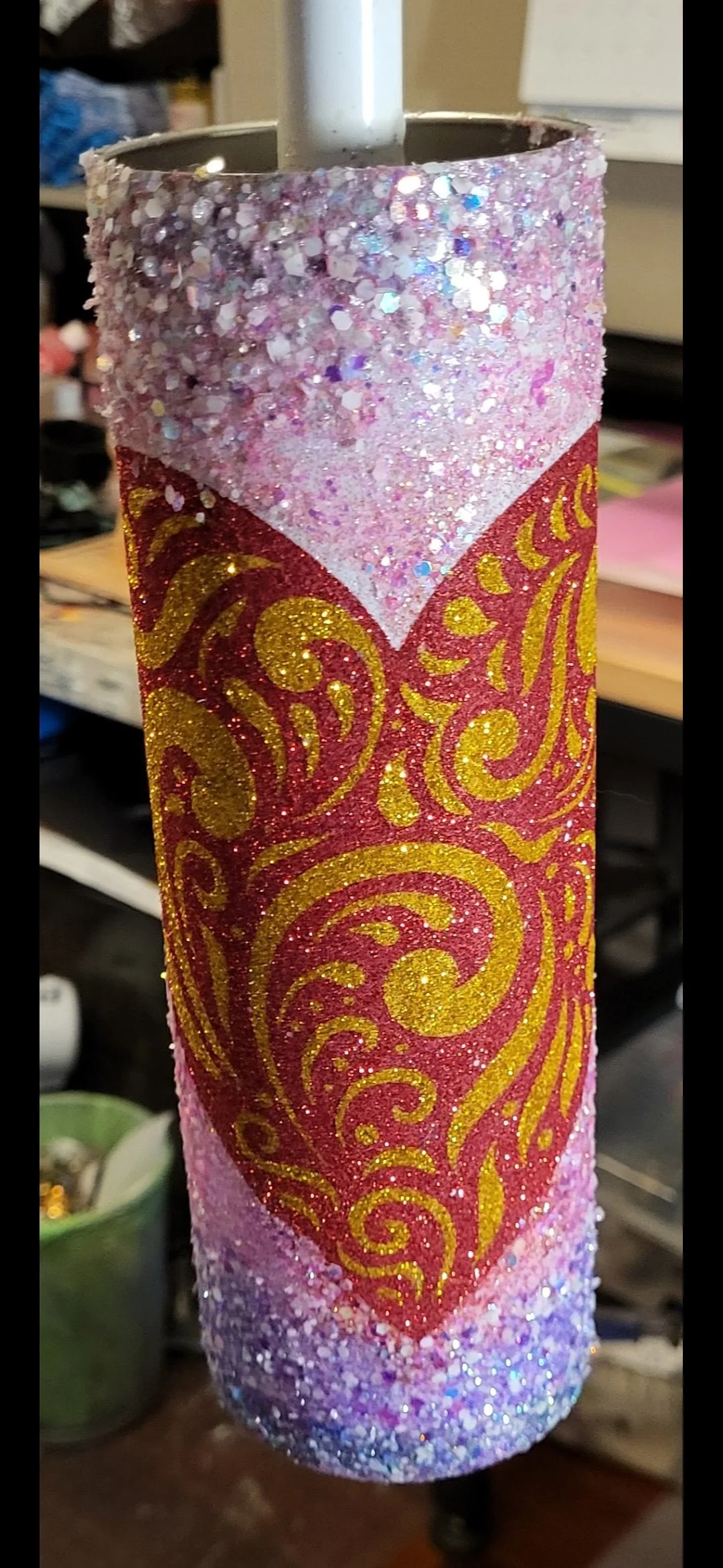 A cylindrical craft object decorated with pink, gold, and purple glitter, featuring a heart shape with gold swirls, topped with a white stick.