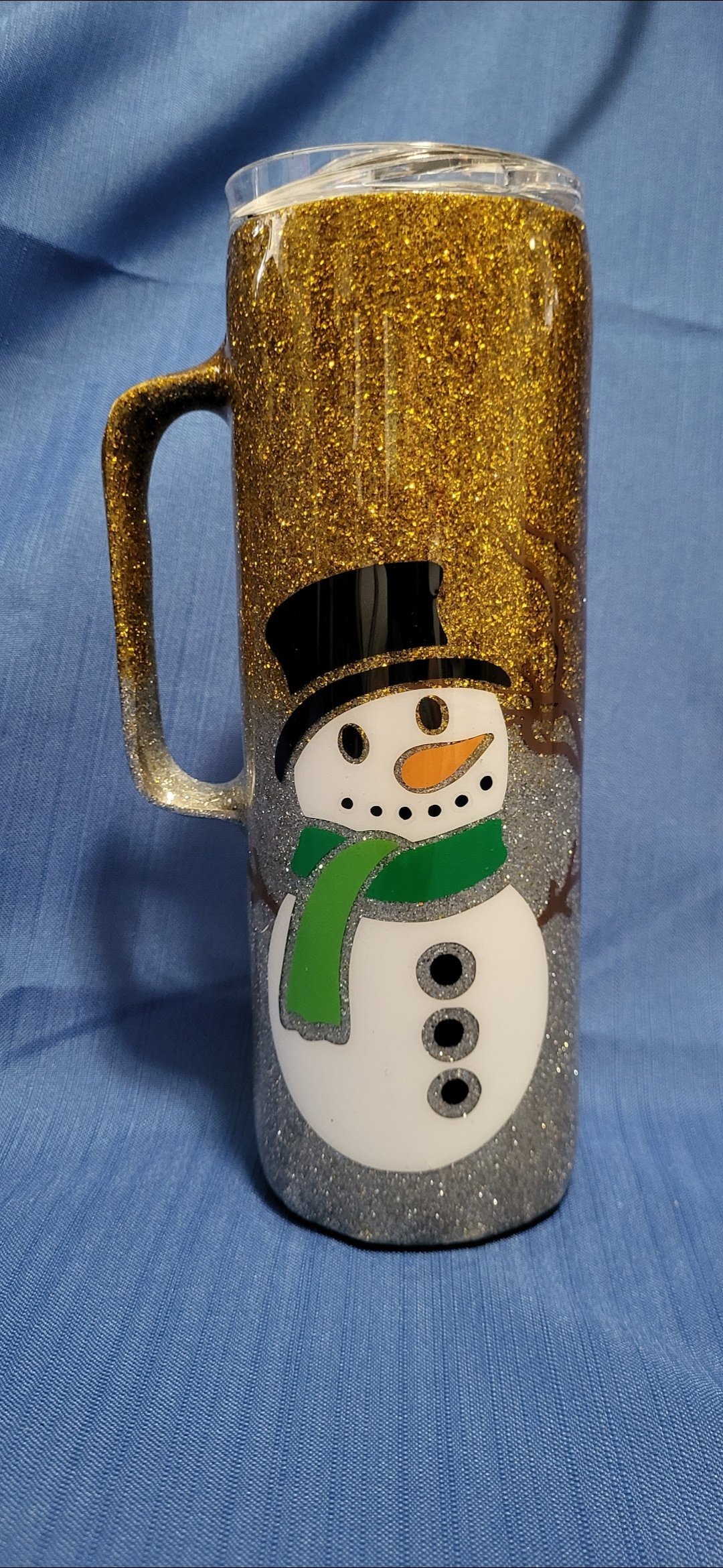 A tall, clear plastic mug with a snowman design, filled with a sparkling gold-colored beverage and topped with a transparent lid, set on a blue fabric background.