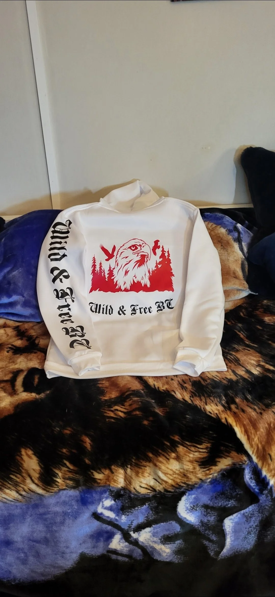 White hoodie with a red eagle graphic and black text reading 'Wild & Free BC' on the front, laid on a blanket with a tiger and wolf print, on a bed with pillows.