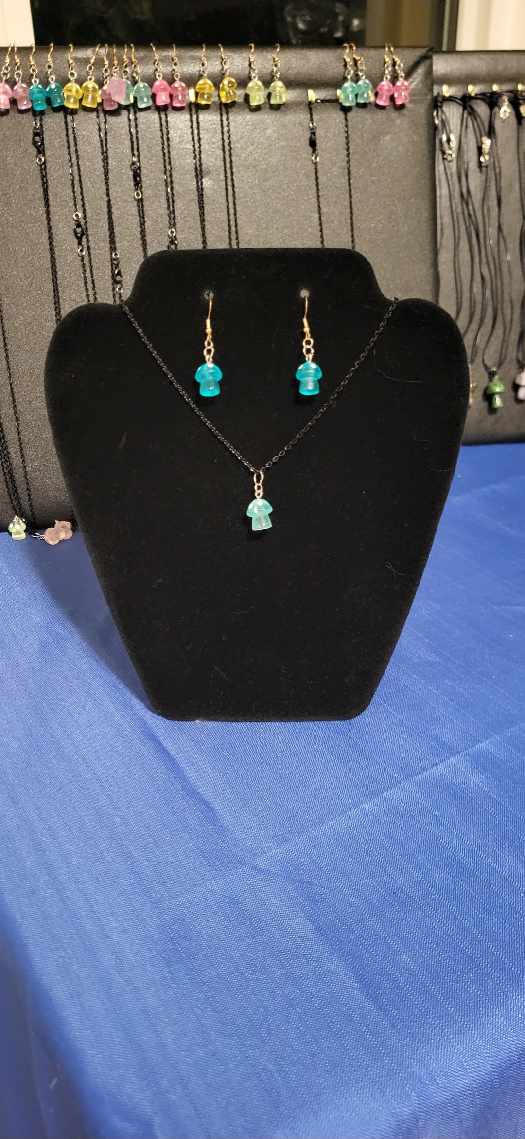 A jewelry display with a black velvet bust showcasing a necklace and matching earrings with blue mushroom-shaped pendants. In the background, there is a display of similar necklaces with various colored mushroom pendants hanging on a gray board.