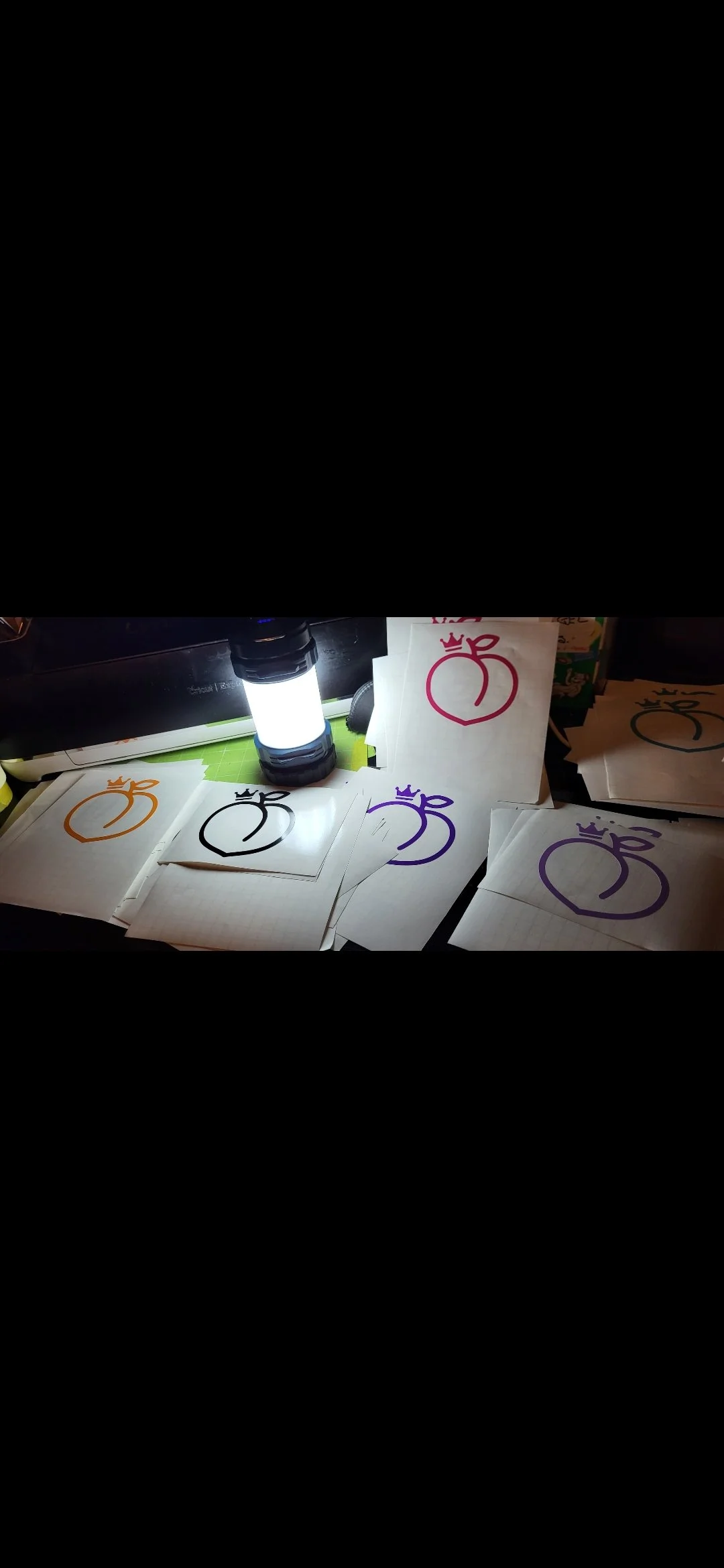 Multiple pieces of paper with colorful crown and fruit-like logo designs, illuminated by a small bright flashlight, on a cluttered desk.