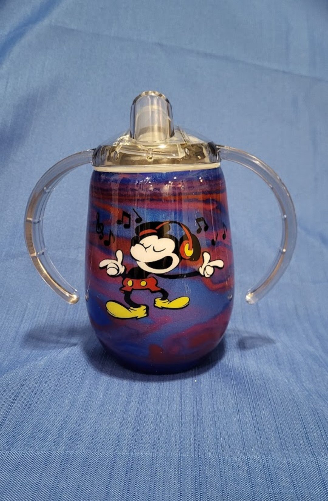 A clear plastic sippy cup with a Mickey Mouse design and colorful swirl background, featuring Mickey Mouse wearing headphones and surrounded by musical notes, placed on a blue fabric surface.
