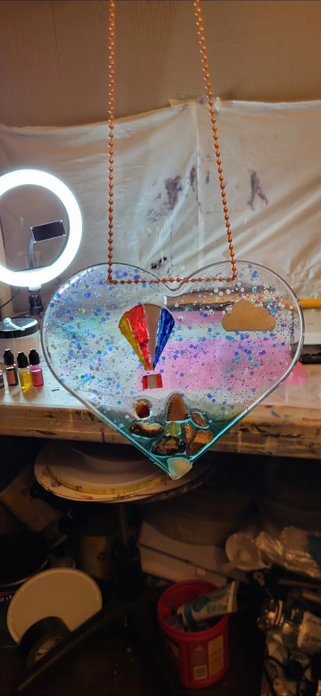 A decorated glass heart with painted hot air balloon and cloud, hanging from an orange beaded chain. The background shows a cluttered workspace with a ring light, bottles of paint, and various art supplies.