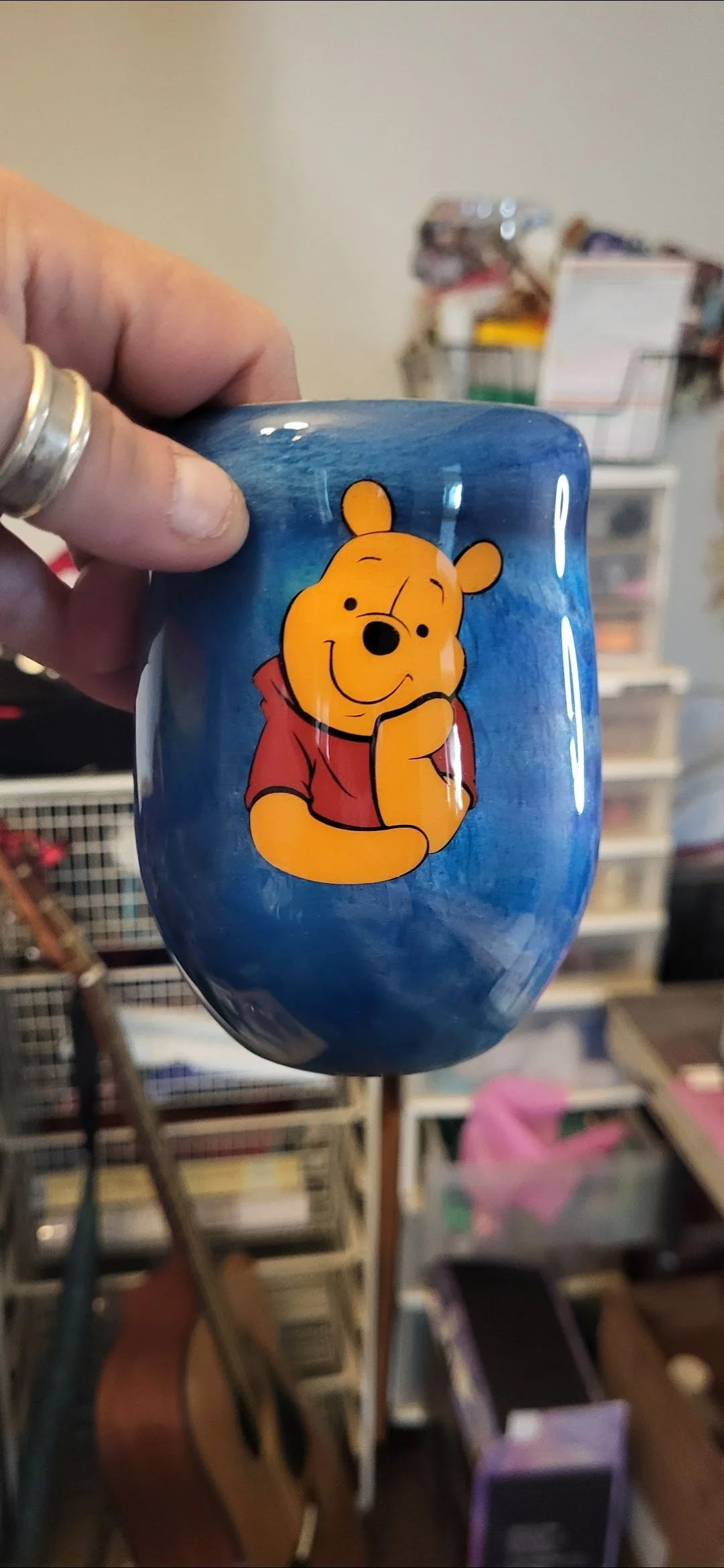 A hand holding a blue ceramic mug with a Winnie the Pooh illustration on it, in a room with various storage containers and objects in the background.