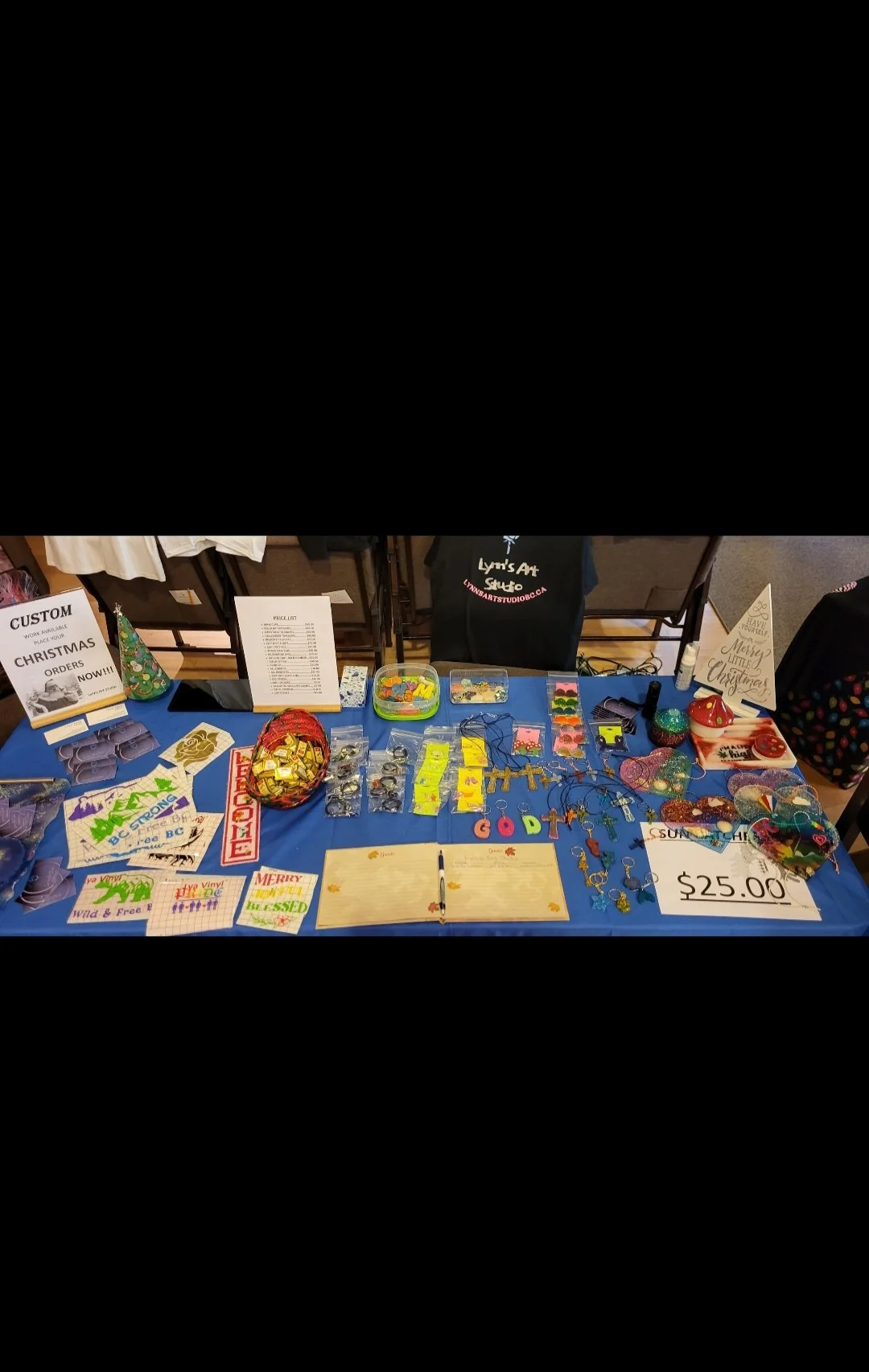Holiday-themed jewelry and accessories on a table for sale at a craft fair, priced at $25.00, with signs and promotional materials.