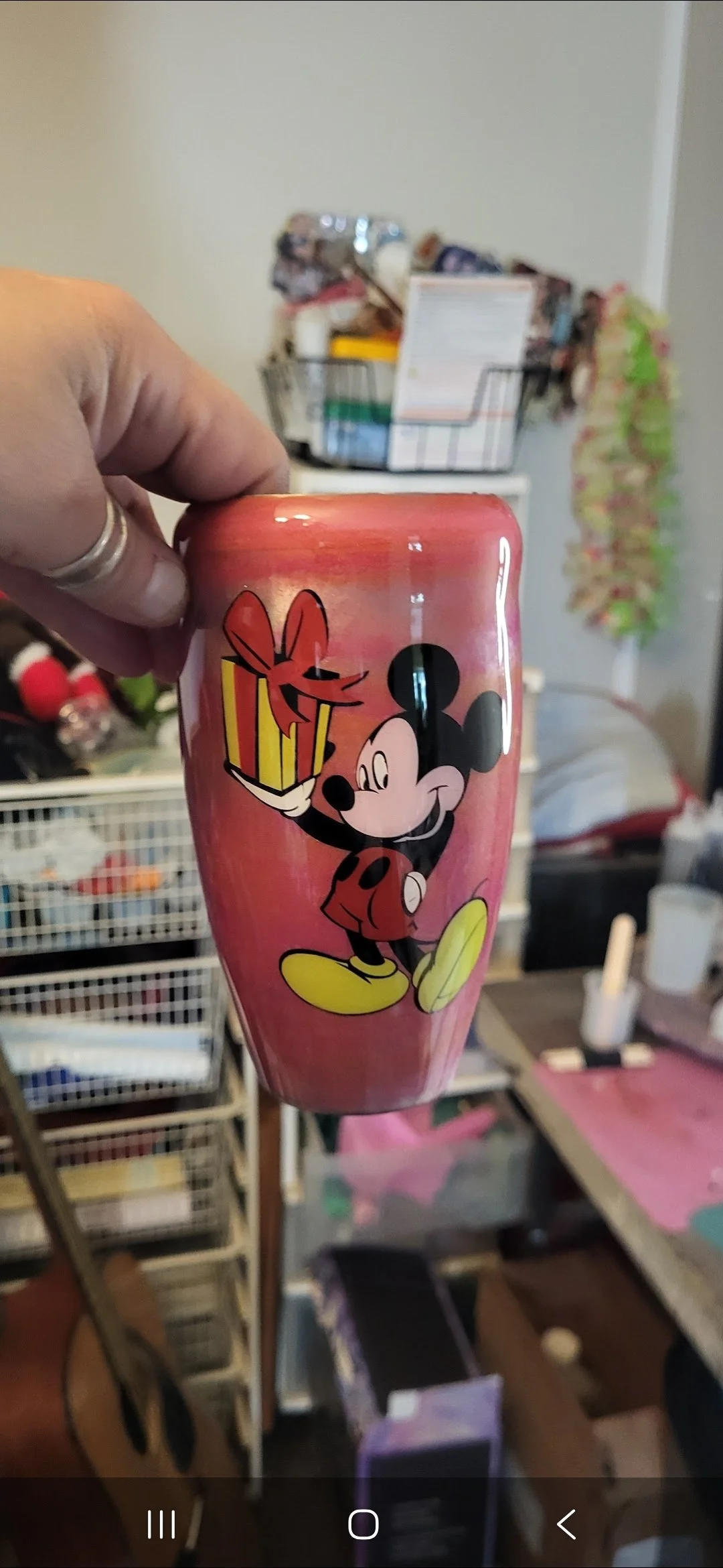 A hand holding a pink Mickey Mouse themed cup with a gift box graphic. In the background, there are various household items and storage containers.