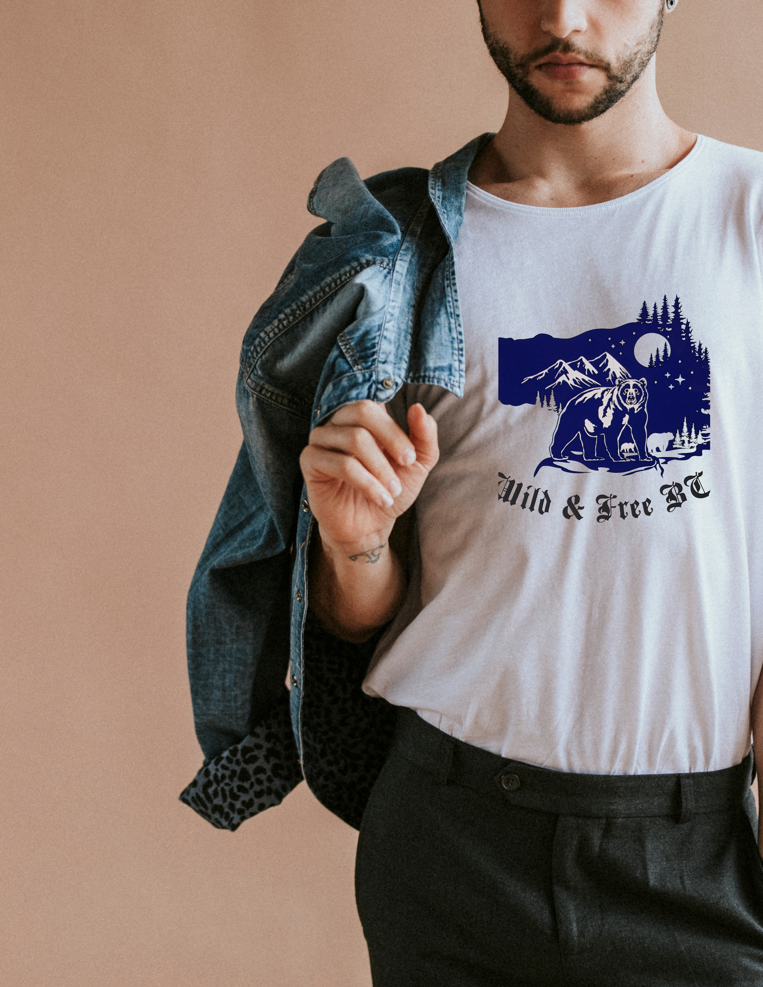 A person wearing a white T-shirt with a forest and mountain design and the words 'Wild & Free BC,' holding a denim jacket over their shoulder.