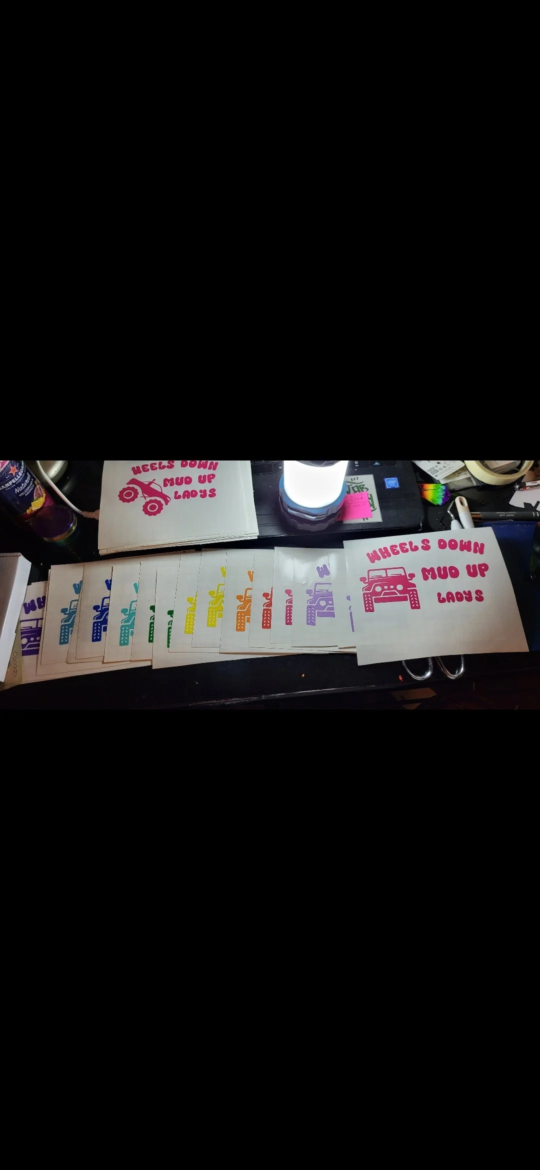 Colorful car-themed stickers with the text "Wheels Down Mud Up Lady's" arranged on a cluttered desk.