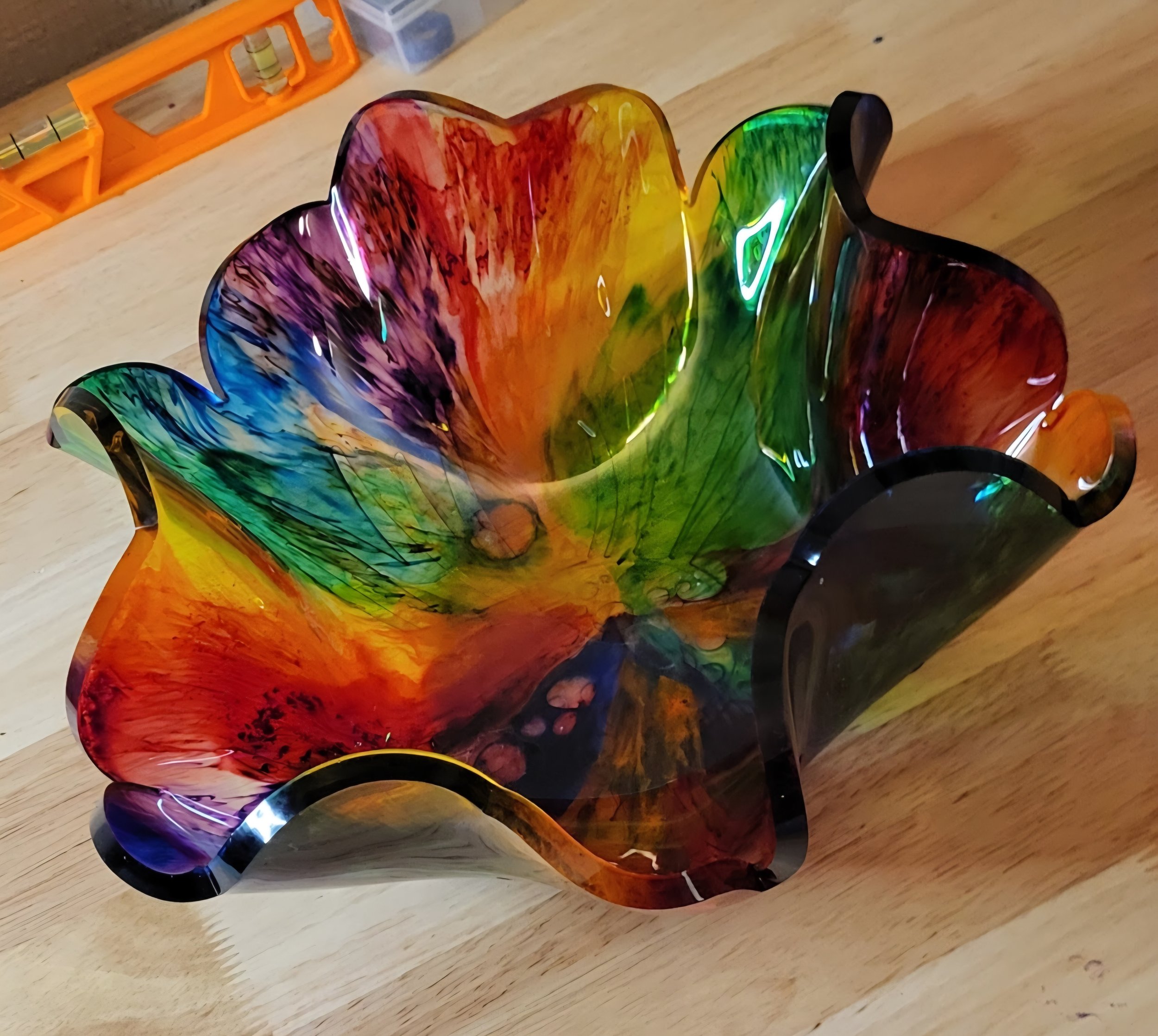 A colorful, hand-blown glass bowl with a ruffled edge, featuring swirls of red, orange, yellow, green, blue, and purple, sitting on a wooden surface.