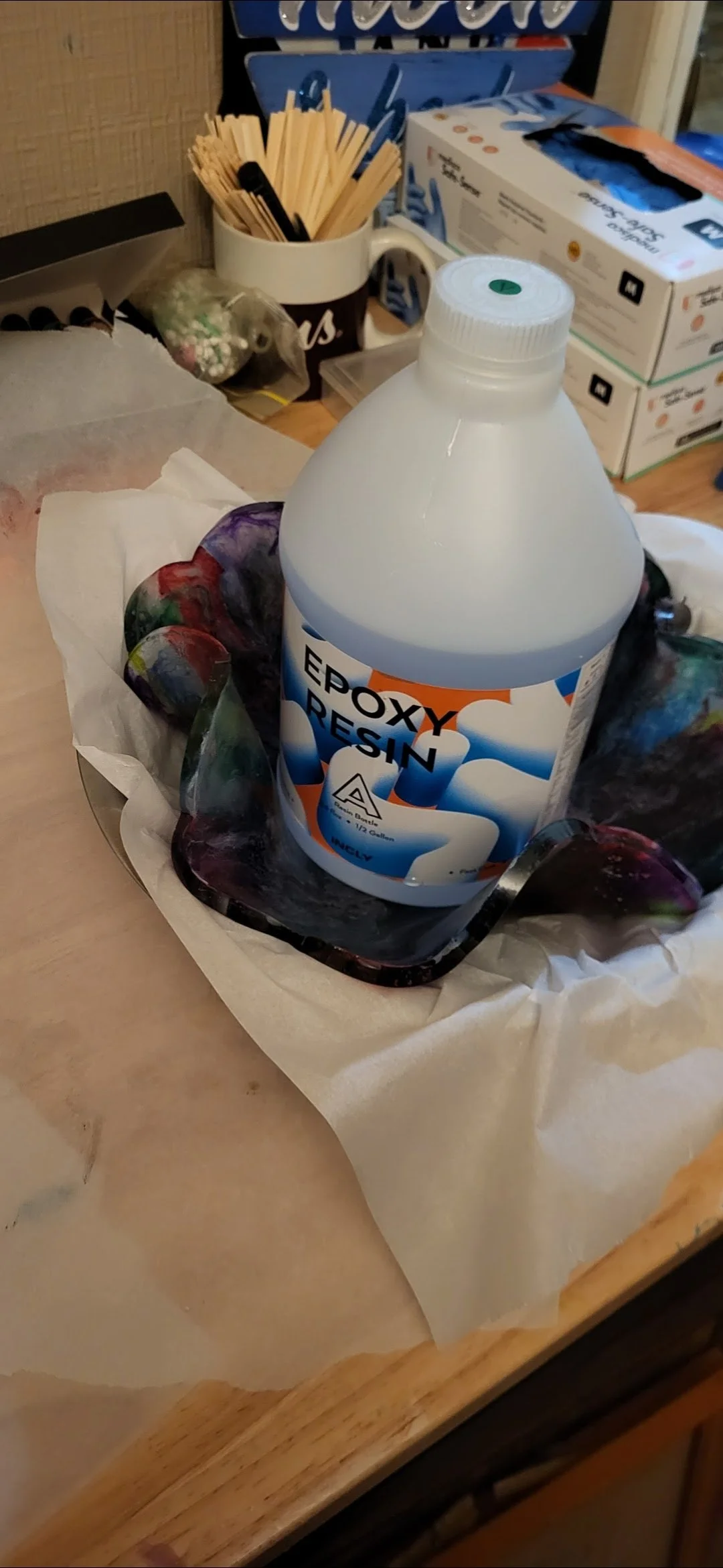 A bottle of epoxy resin on a countertop, surrounded by colorful glass pieces and protective paper, with various supplies in the background.