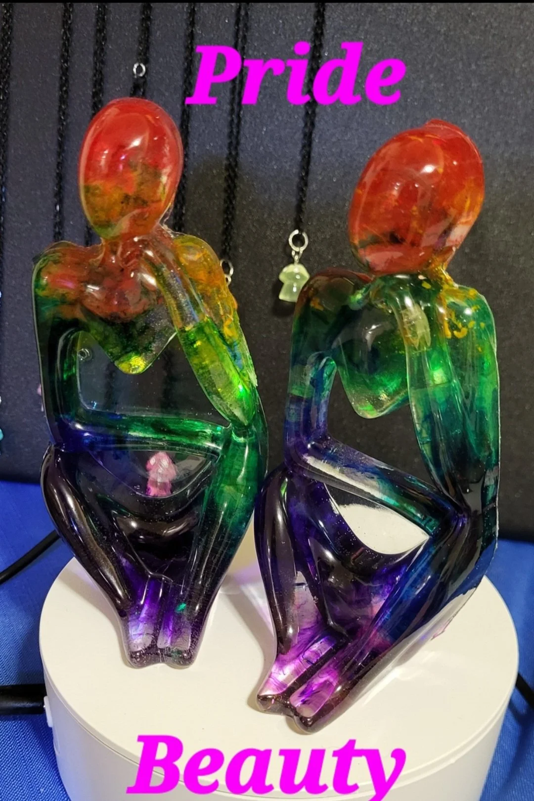 Two colorful glass art sculptures of abstract female figures, one with a pink head and green and purple body, and the other with an orange head and green and purple body, placed on a white pedestal against a black background. The words "Pride" and "B