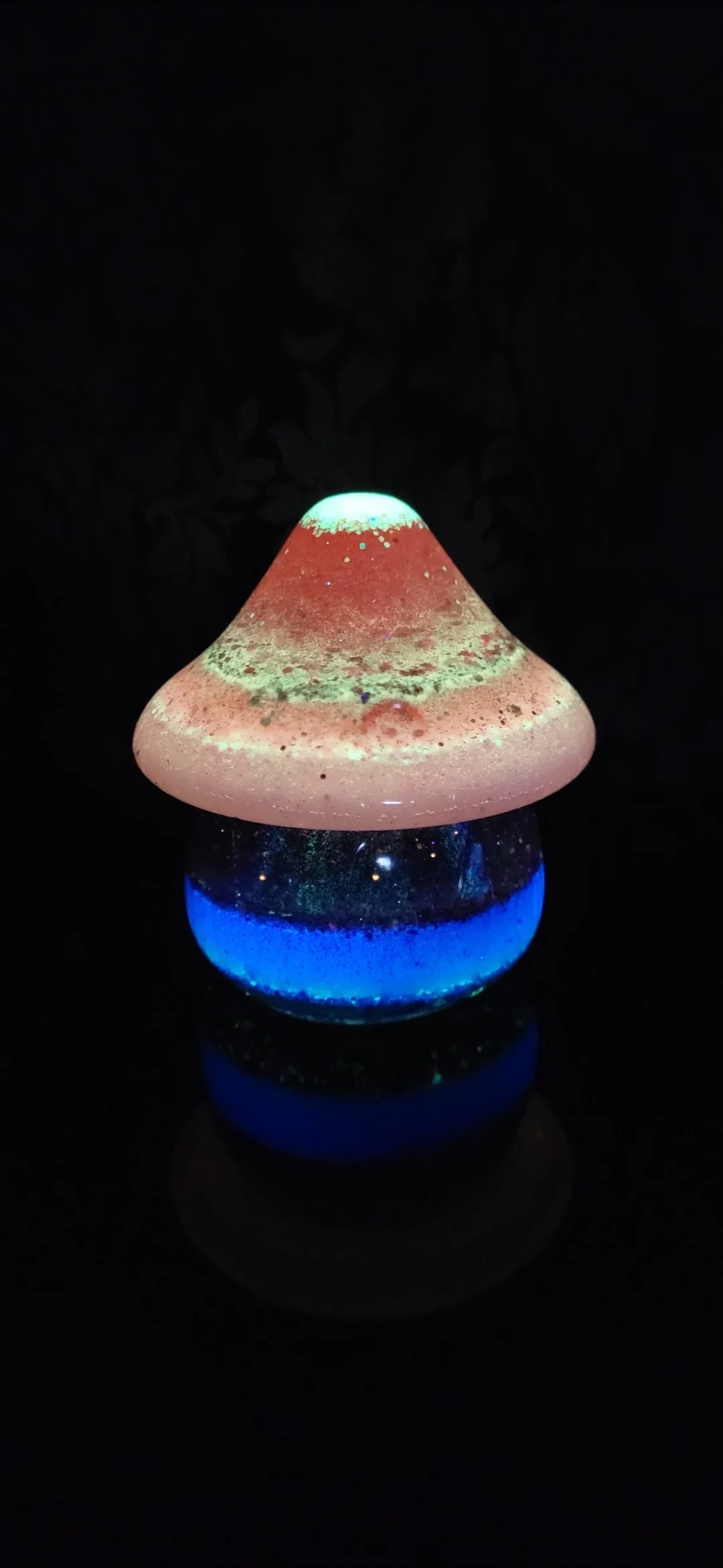 A glass mushroom-shaped lamp with a glowing white to red cap and a glowing blue base, emitting colorful light in a dark setting.