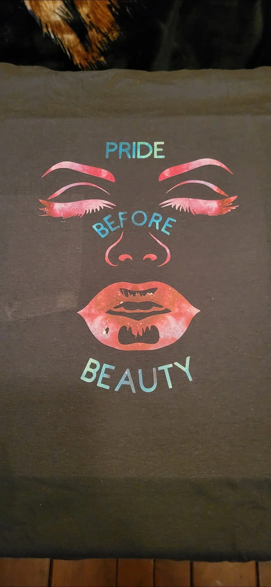 Illustration of closed eyes with long eyelashes, a nose, and lips, with colorful text saying "PRIDE BEFORE BEAUTY" on a dark background.