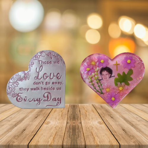 Two heart-shaped memorial stones on a wooden table, one with an embroidered quote about love and the other with a photo of a woman surrounded by flowers and a cross, with a blurred warm background.