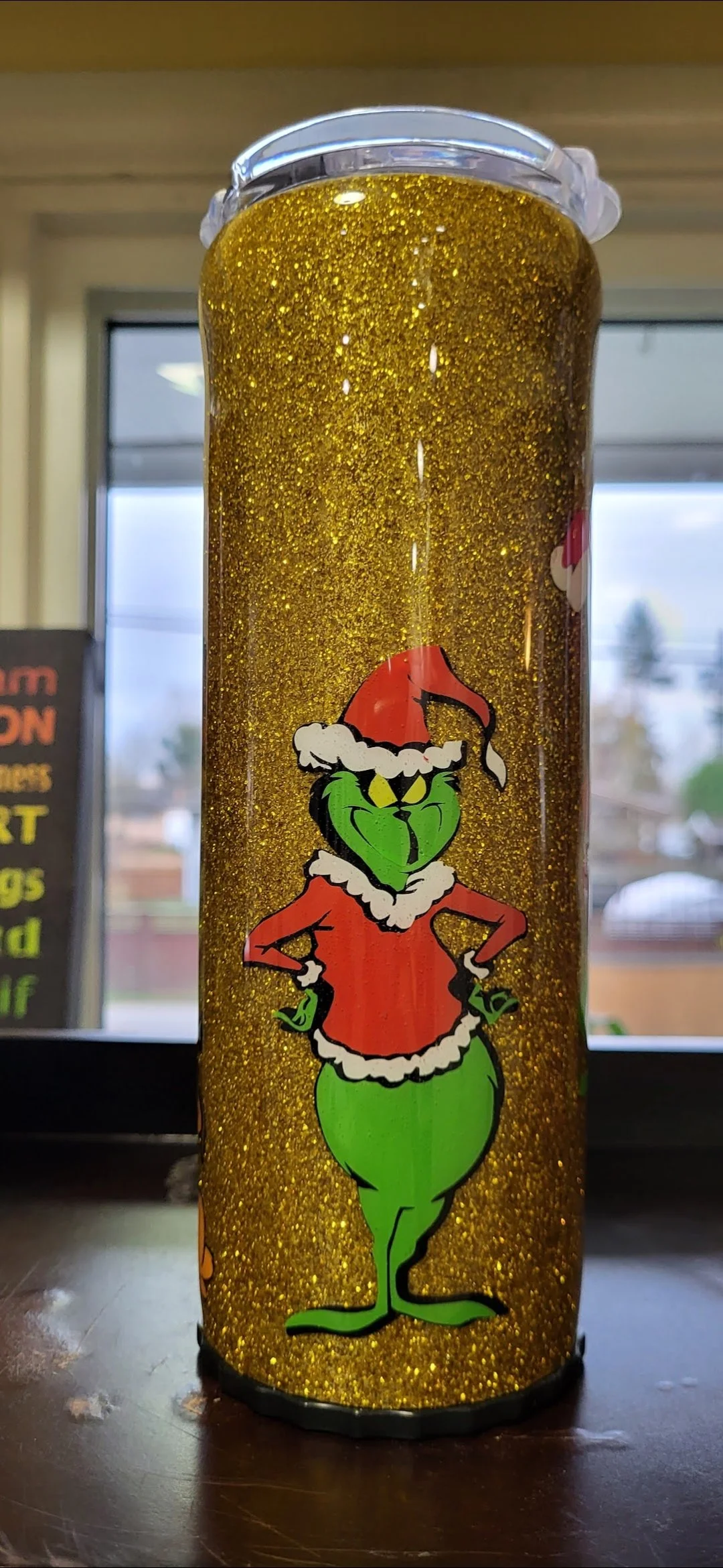 A tall cylindrical holiday-themed tumbler with a gold glitter background and a cartoon character of the Grinch dressed as Santa Claus.