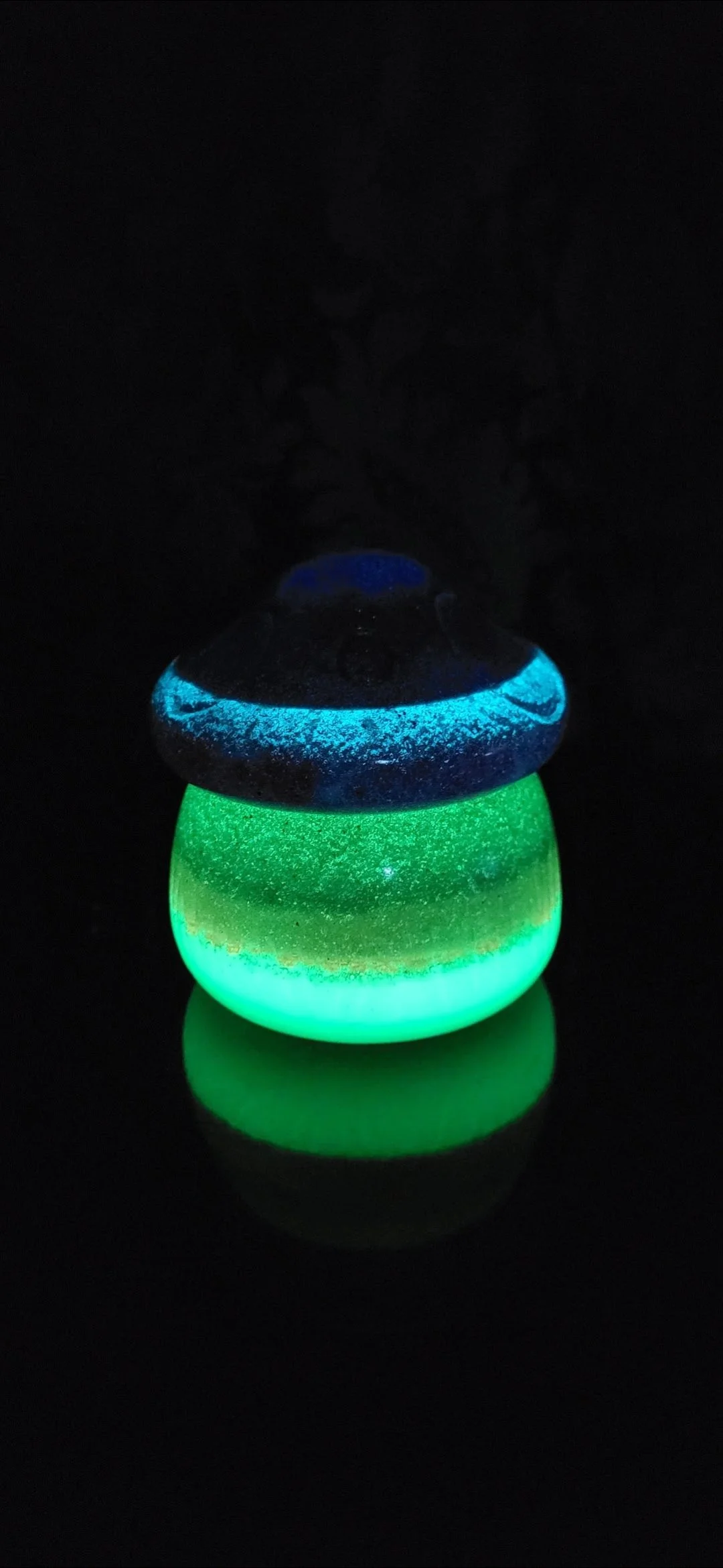 Glow-in-the-dark mushroom-shaped object with a green base and blue and black cap, reflecting on a black surface.