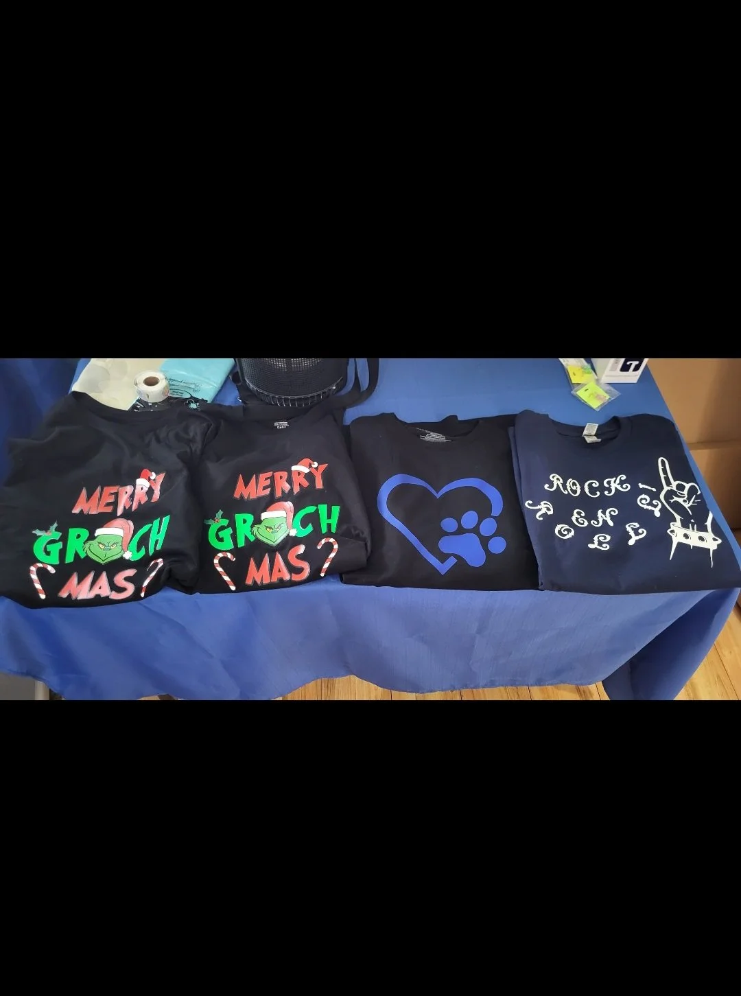 Display of four T-shirts on a table, two with Christmas-themed graphics and the words "Merry Grolchmas," one with a blue Paw Patrol logo, and one with a rock and roll design.