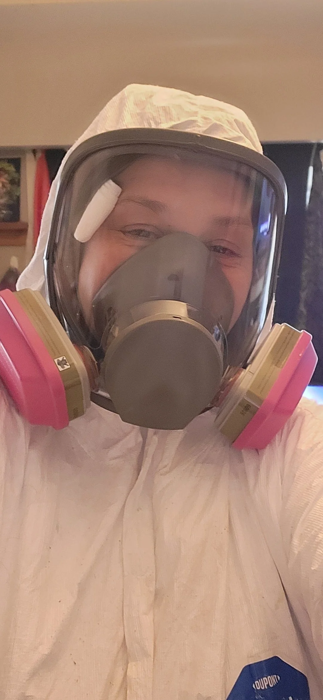 Person wearing a full protective suit, including a respirator mask with pink filters, goggles, and a hood. The individual is inside a room.