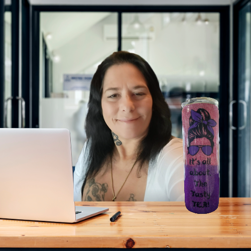 Woman with dark hair, tattoos, and necklace sitting at a wooden table with a laptop and a purple can featuring a graphic of a girl with sunglasses and the text 'It's all about the rest, TERY' in front of her.